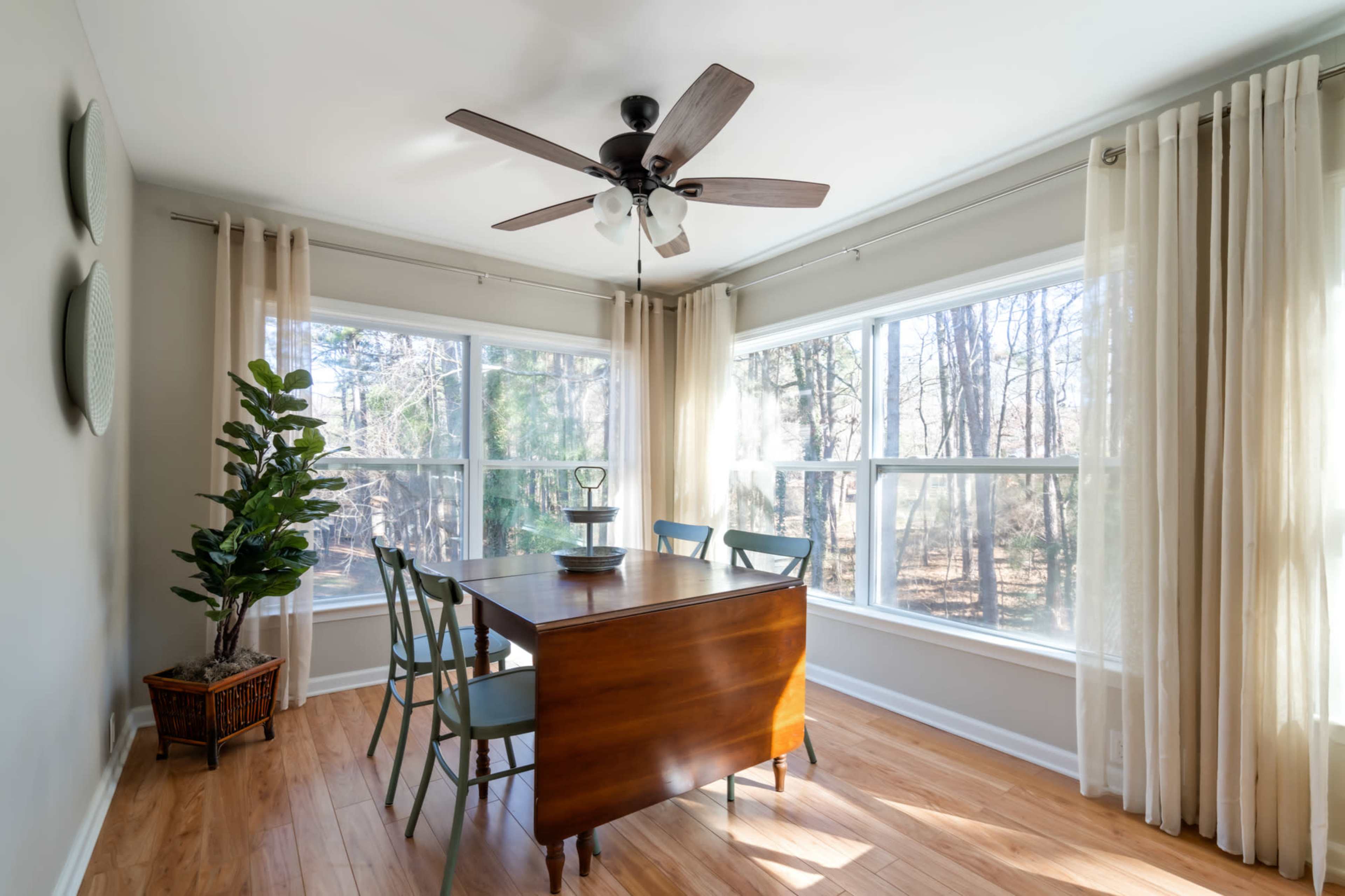 A dining room features a wooden table surrounded by green chairs, large windows with sheer curtains, and a ceiling fan, all set against a backdrop of trees.