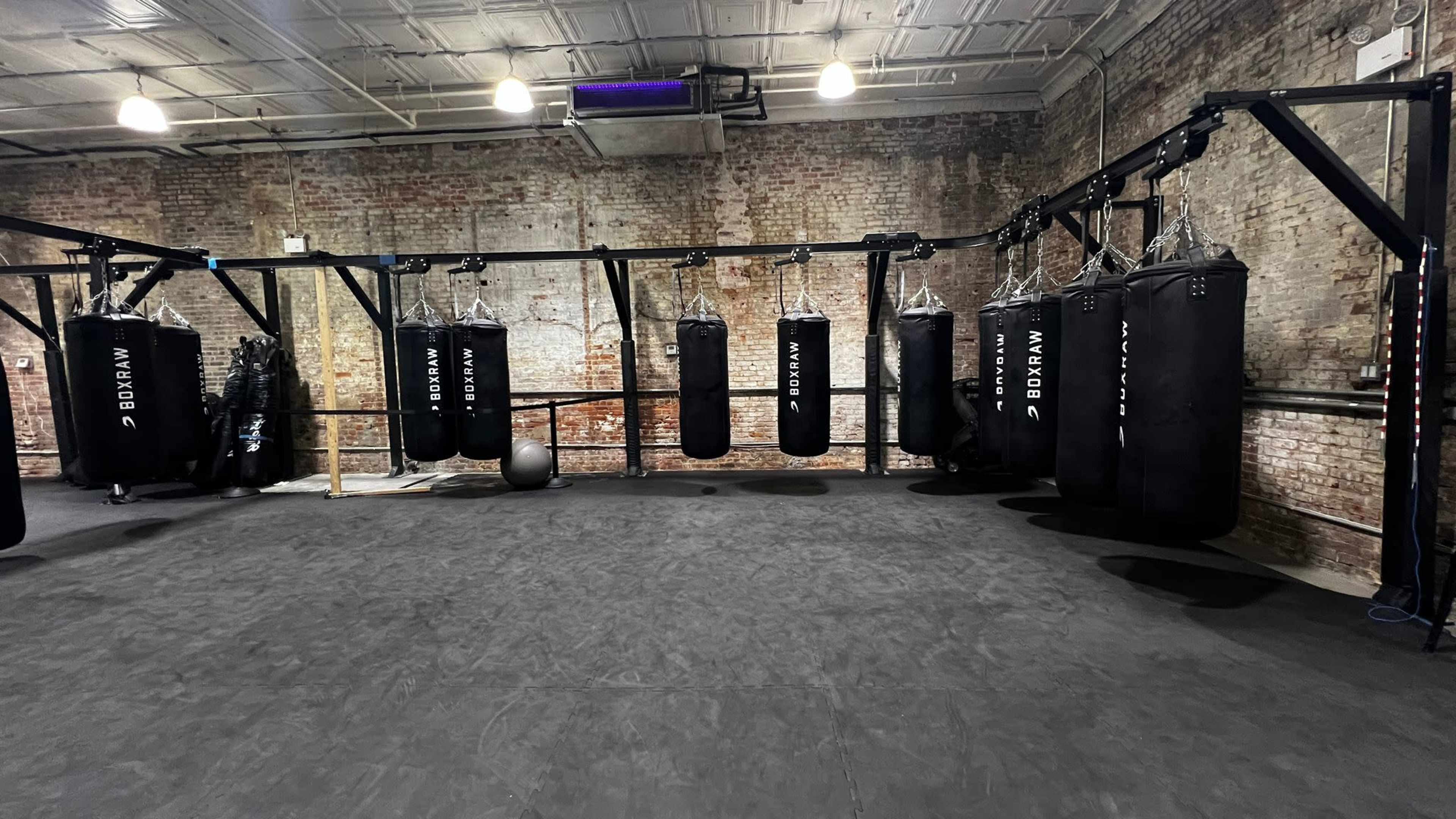 The image shows a gym with several black punching bags suspended from a metal frame against a rustic brick wall.