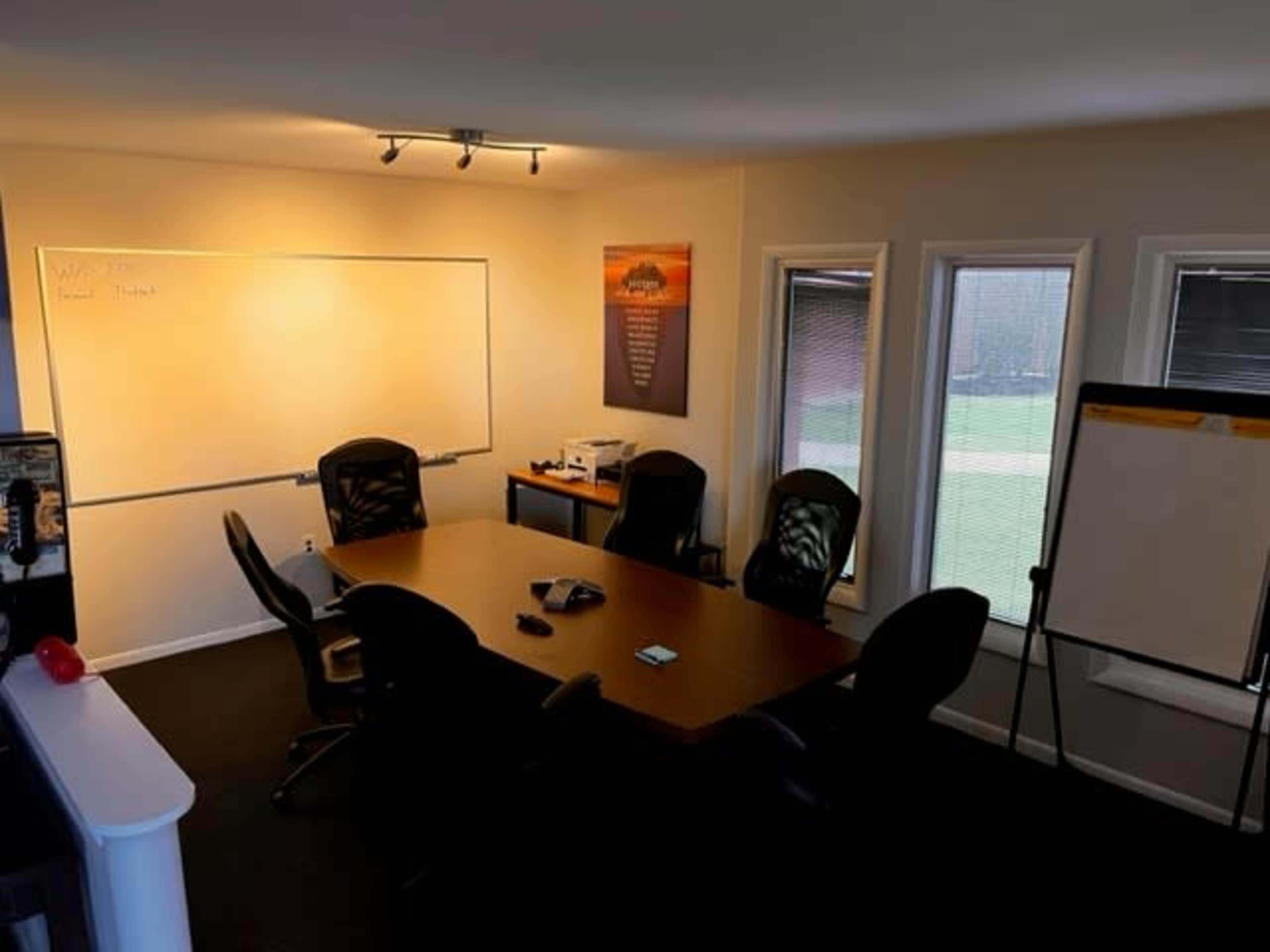 The image shows a meeting room with a large wooden table surrounded by black chairs, a whiteboard on the wall, a flip chart stand, and windows letting in natural light.