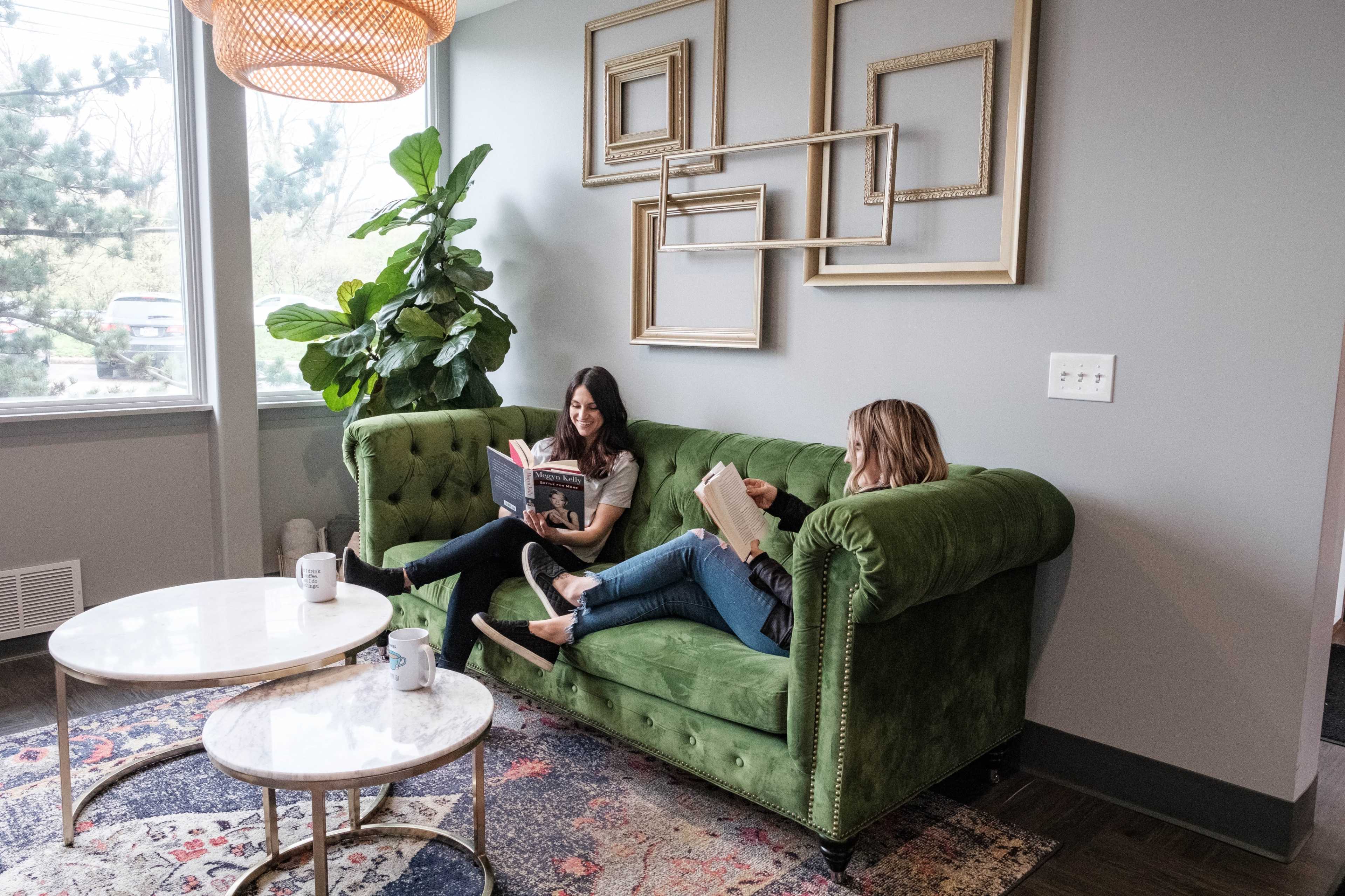 Two women are sitting on a green couch in a bright room, reading books while surrounded by plants and framed art.