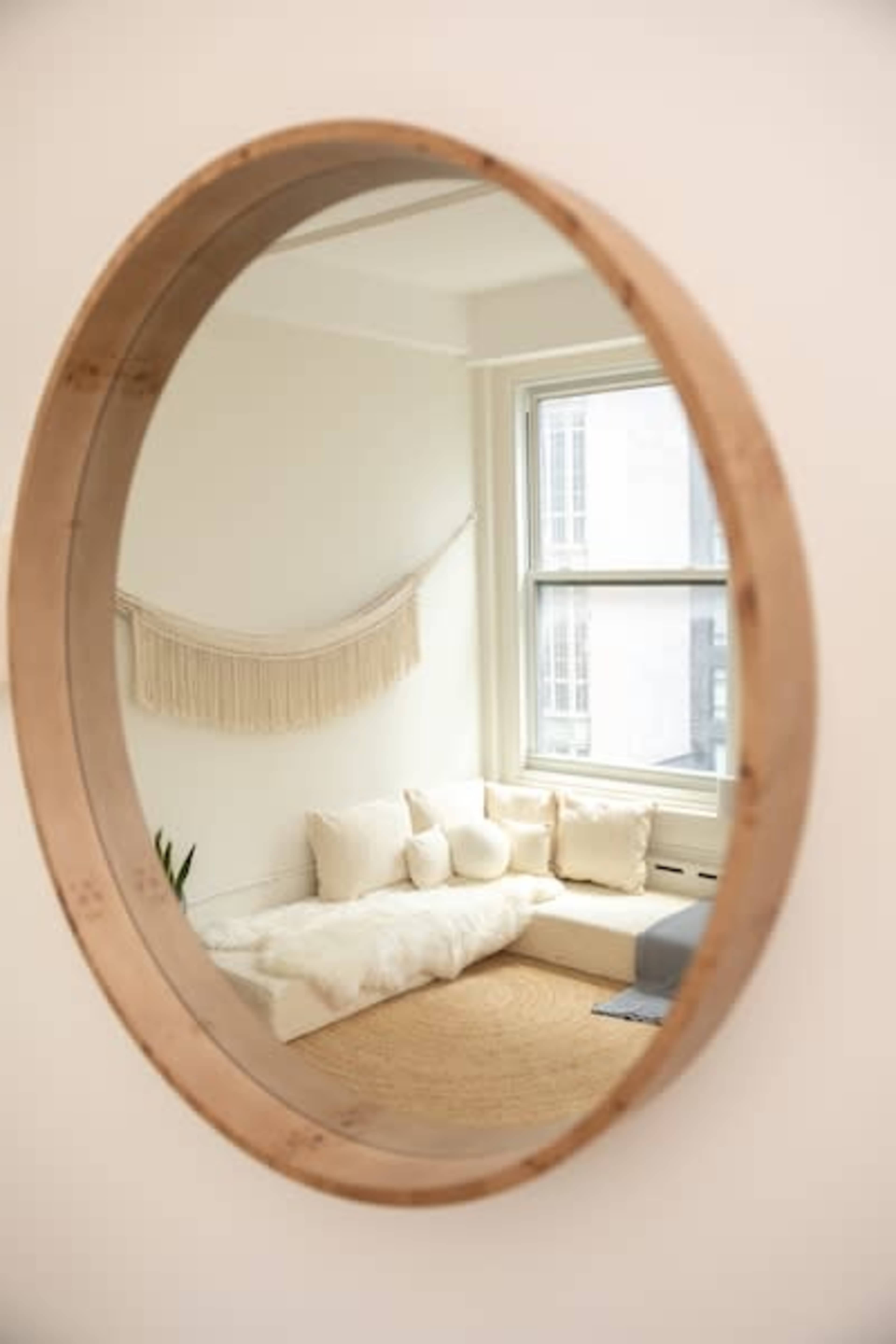 A round wooden mirror reflects a softly lit, minimalist living space featuring a cozy seating area with cushions and a tasseled wall hanging.