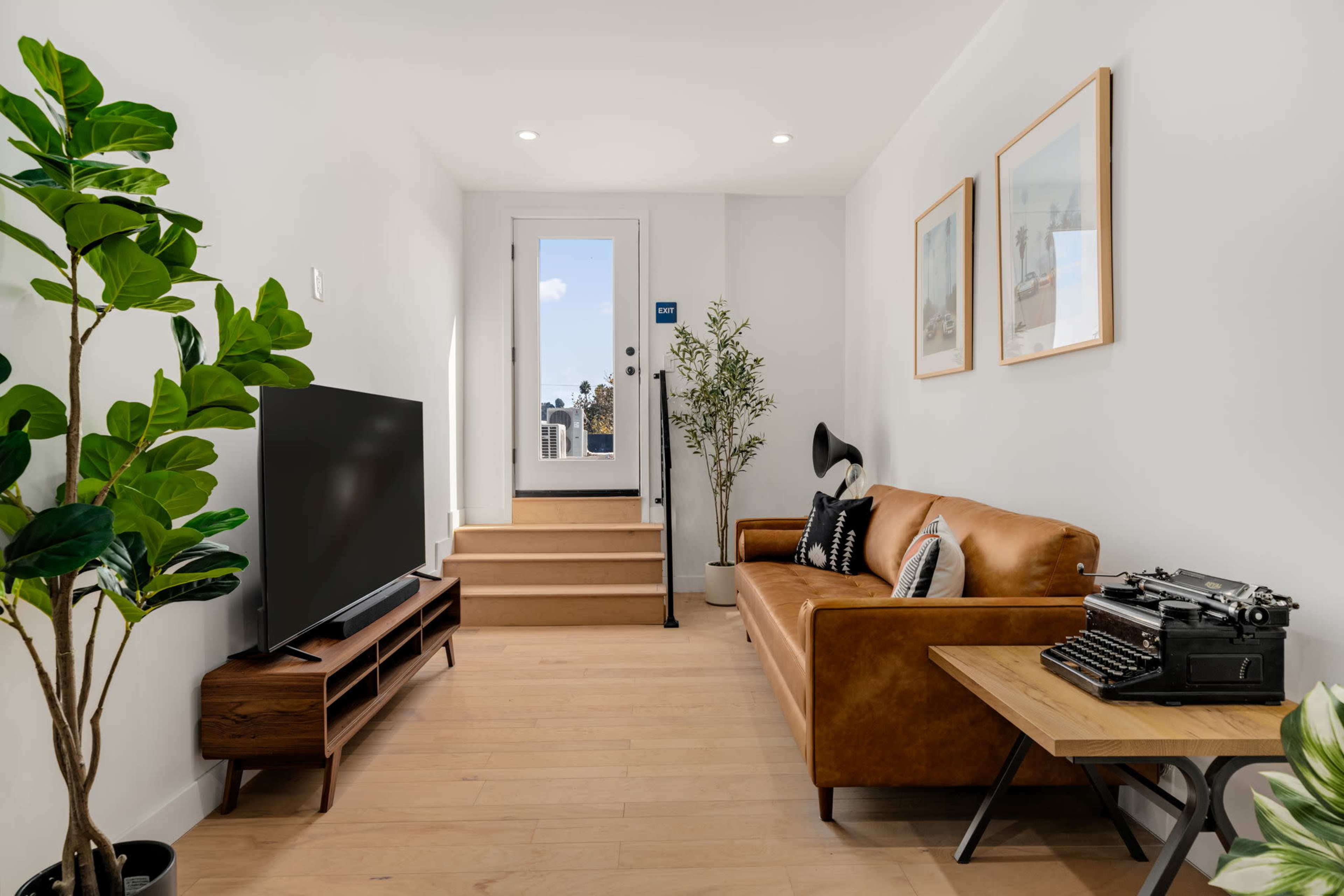A well-lit living room features a brown leather sofa, a wooden TV stand with a black television, a vintage typewriter on a table, and a door leading to a staircase.
