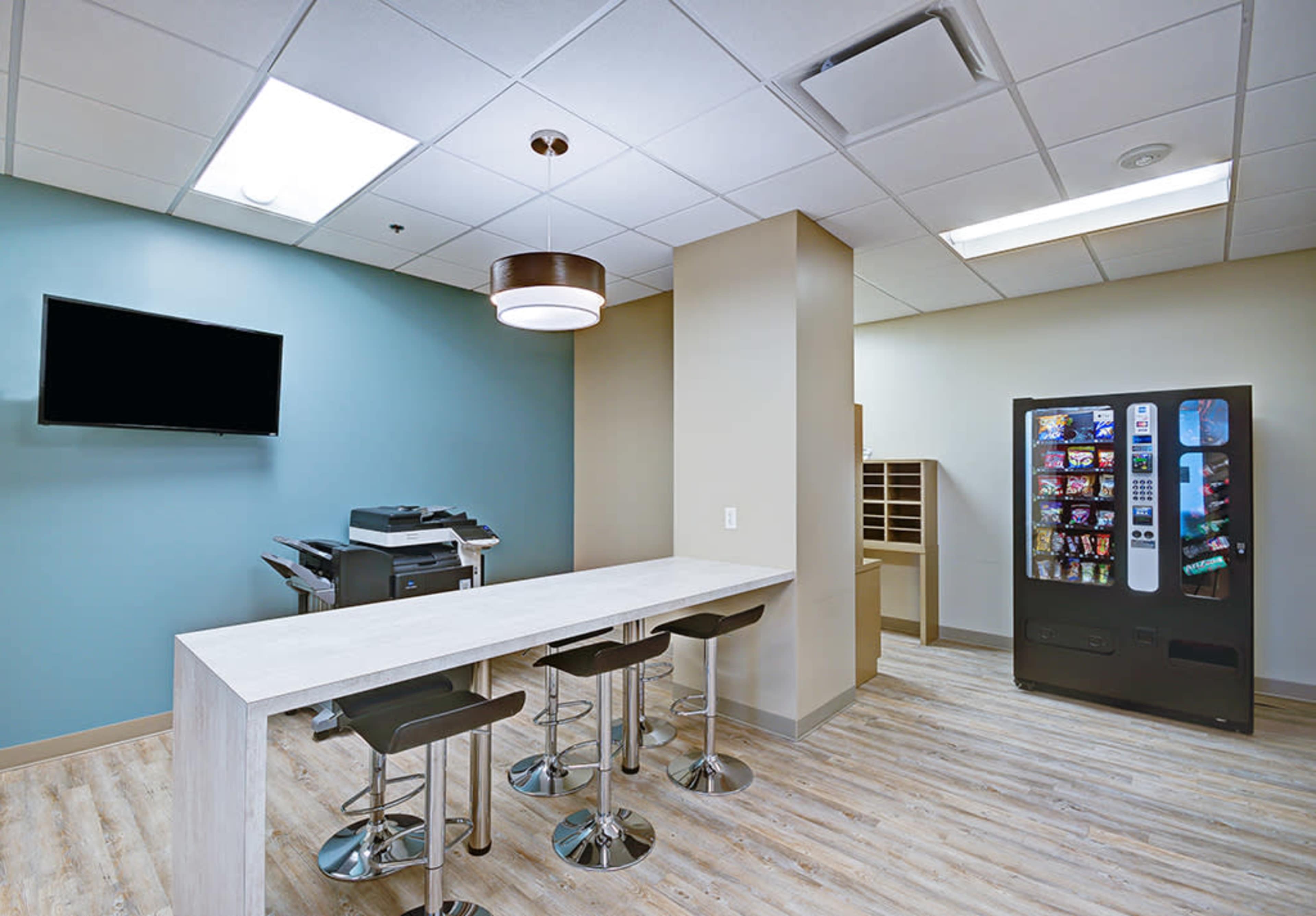 A modern break room with a table and stools, a vending machine, a printer, and a wall-mounted television.