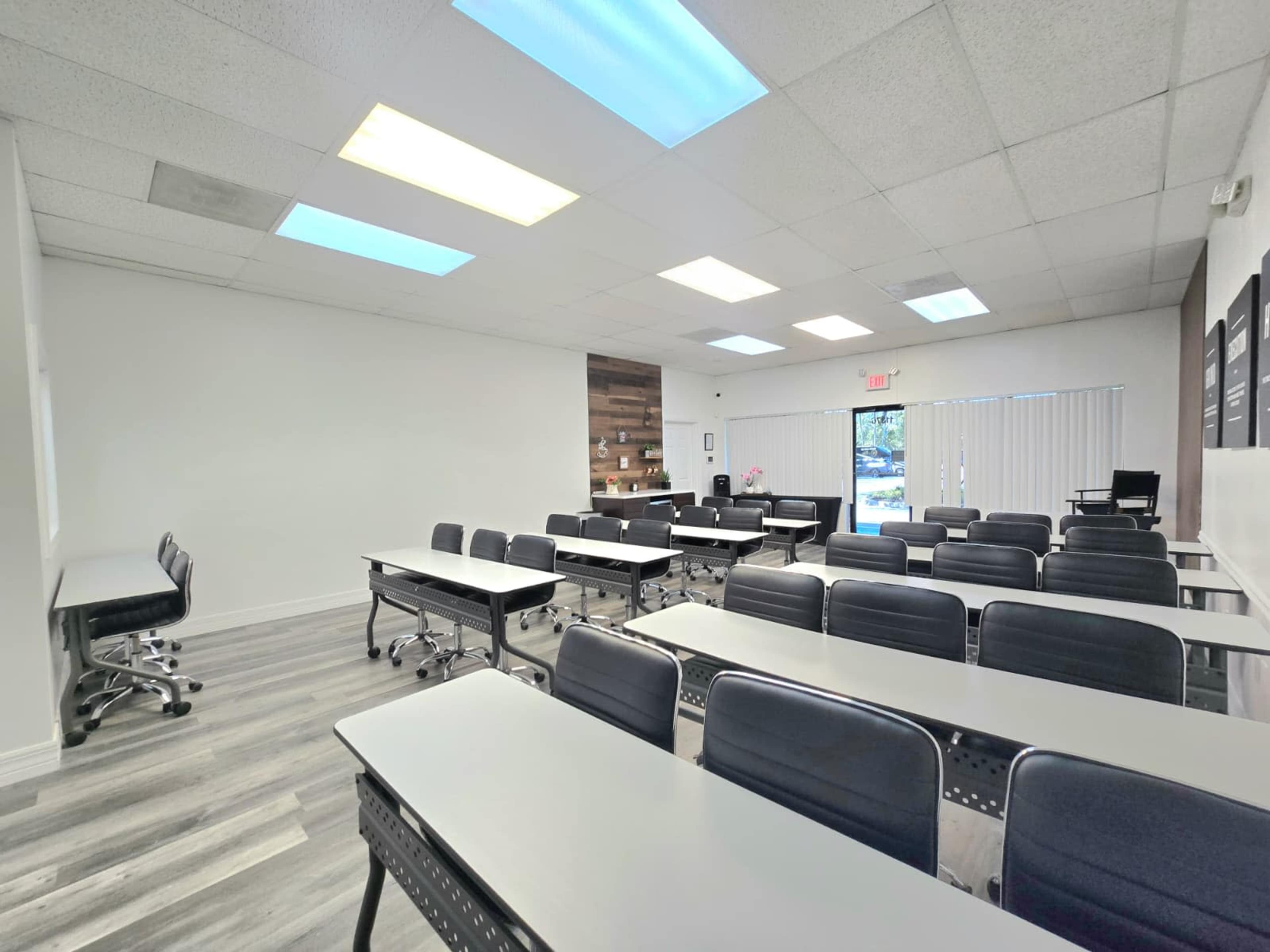 The image shows a modern classroom with several rows of desks and chairs, equipped with windows for natural light and minimalist décor.