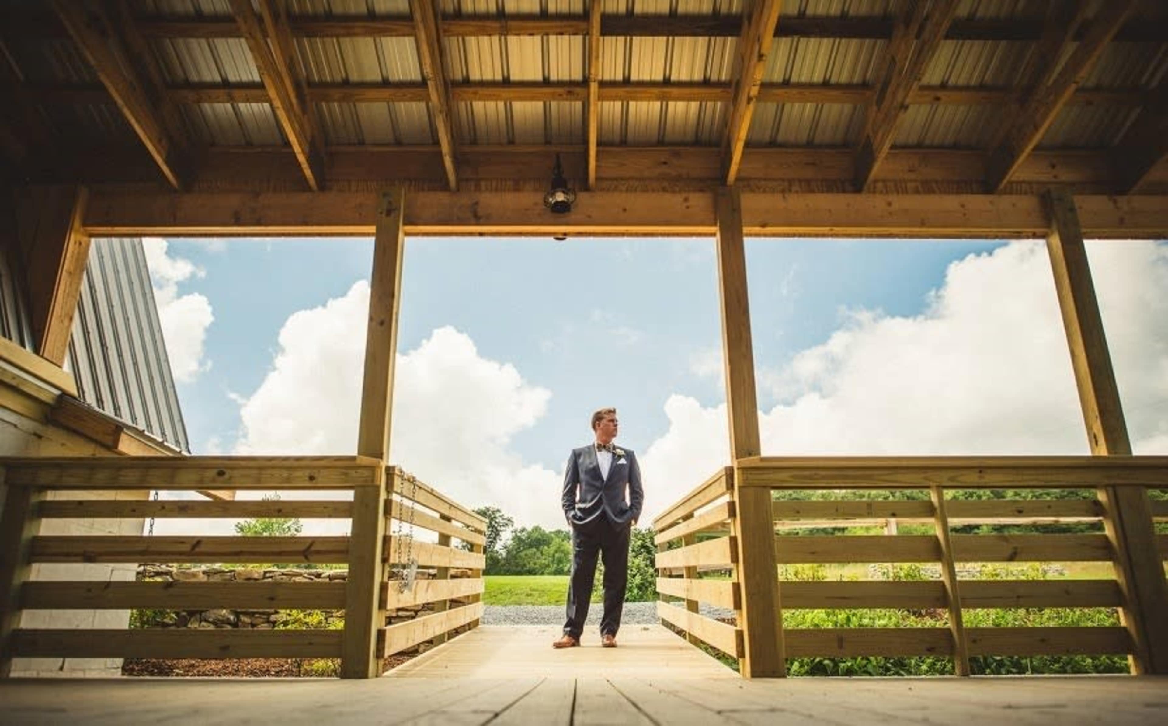 A man in a suit stands on a wooden deck overlooking a landscape with blue skies and clouds.