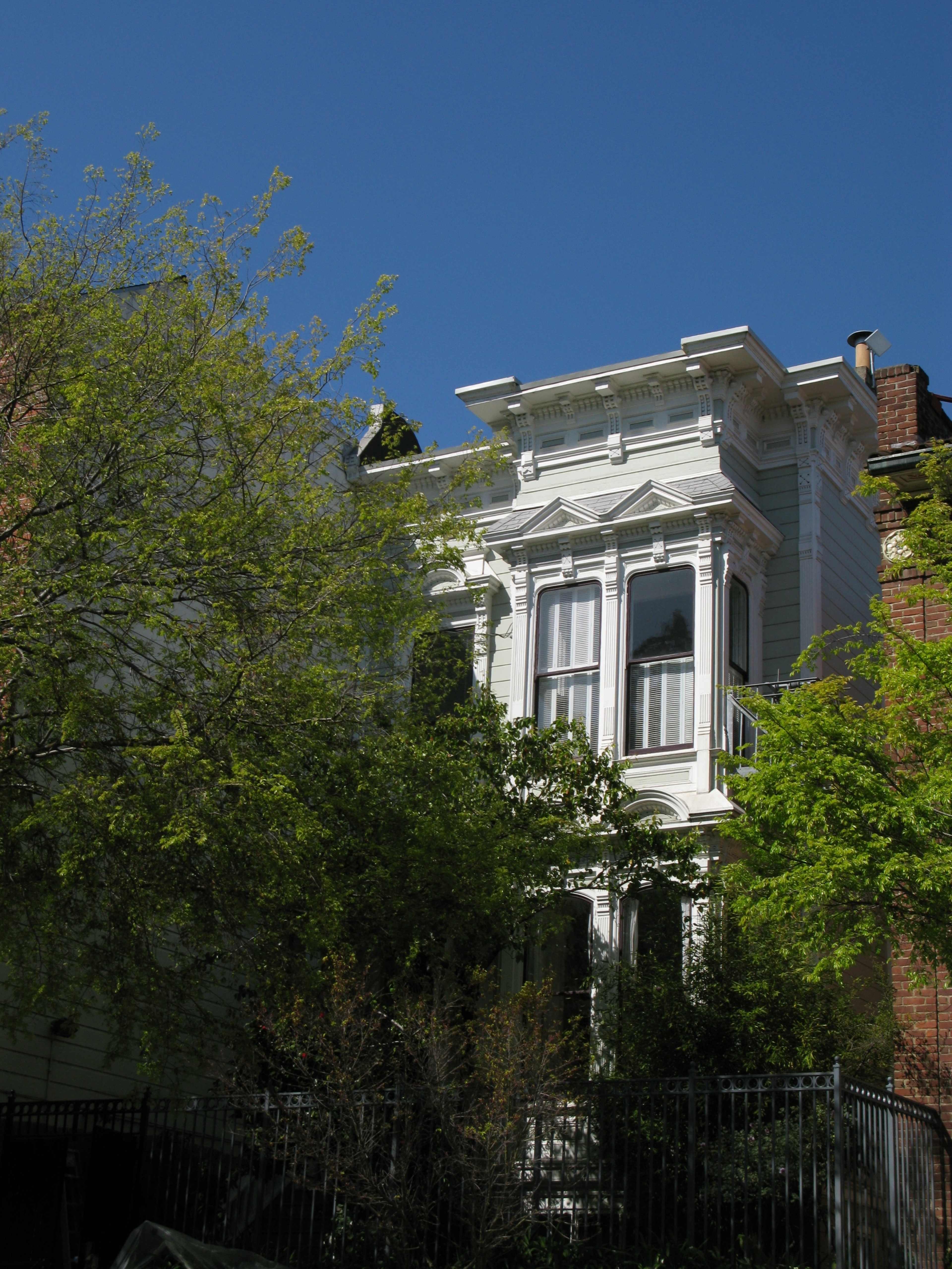 A Victorian-style house with elaborate architectural details is partially obscured by greenery against a clear blue sky.