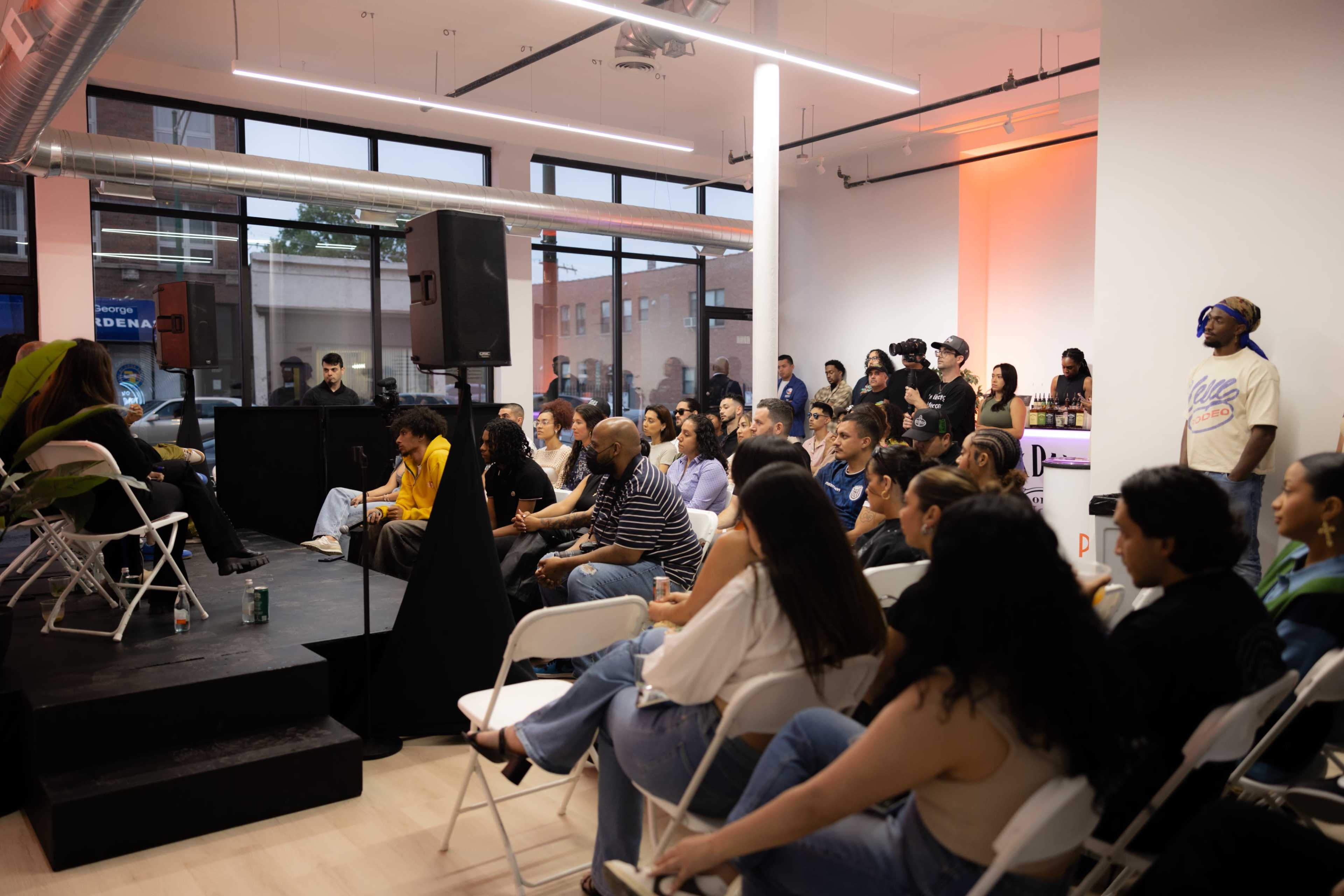 A diverse audience sits in a brightly lit venue, attentively listening to speakers on a stage in front of them.