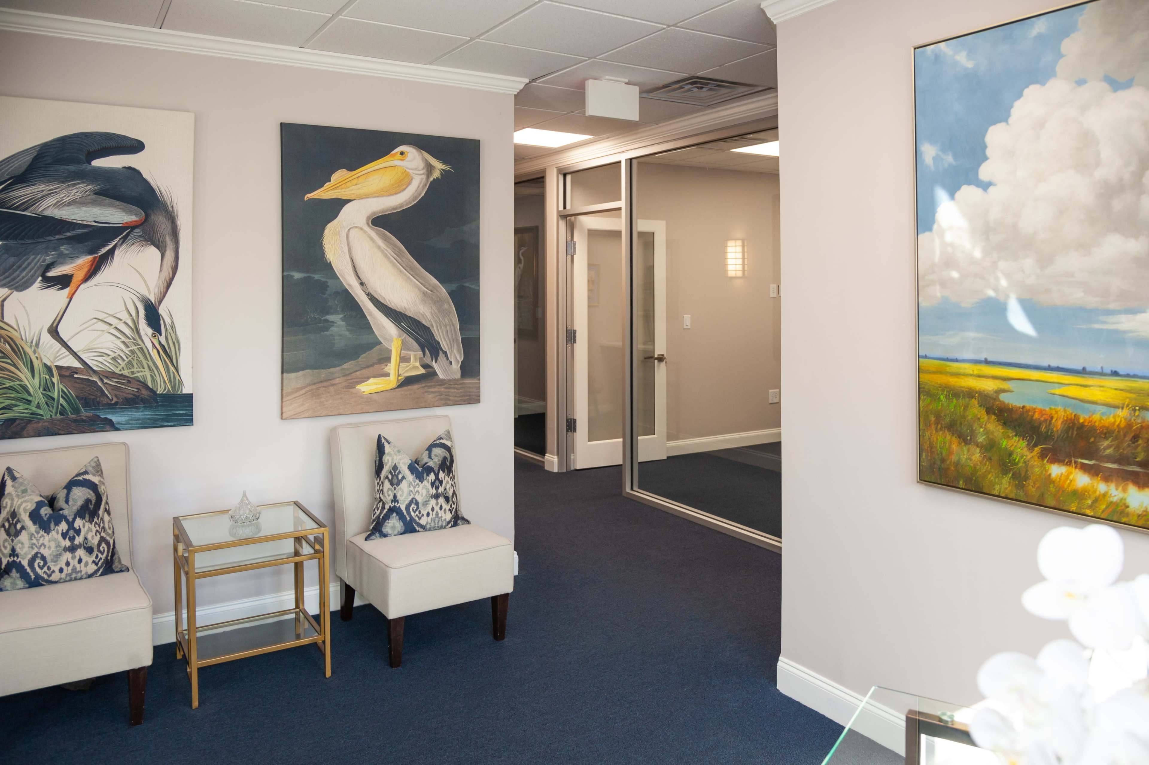 A modern office waiting area featuring two upholstered chairs, a glass-top side table, and large bird-themed artwork on the walls.
