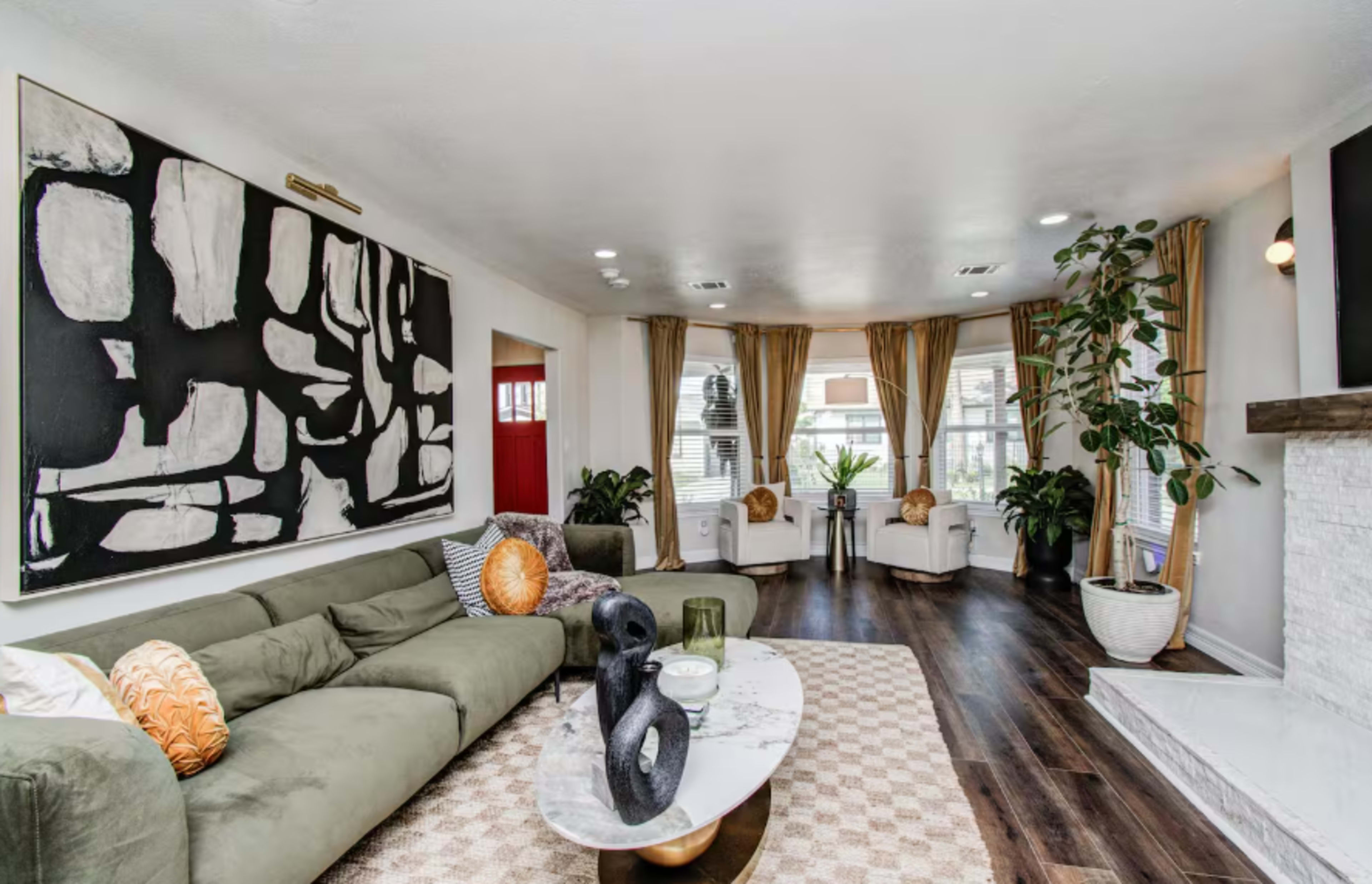 The living room features a green sectional sofa, a marble coffee table, and large windows adorned with curtains, alongside decorative plants and an abstract artwork on the wall.