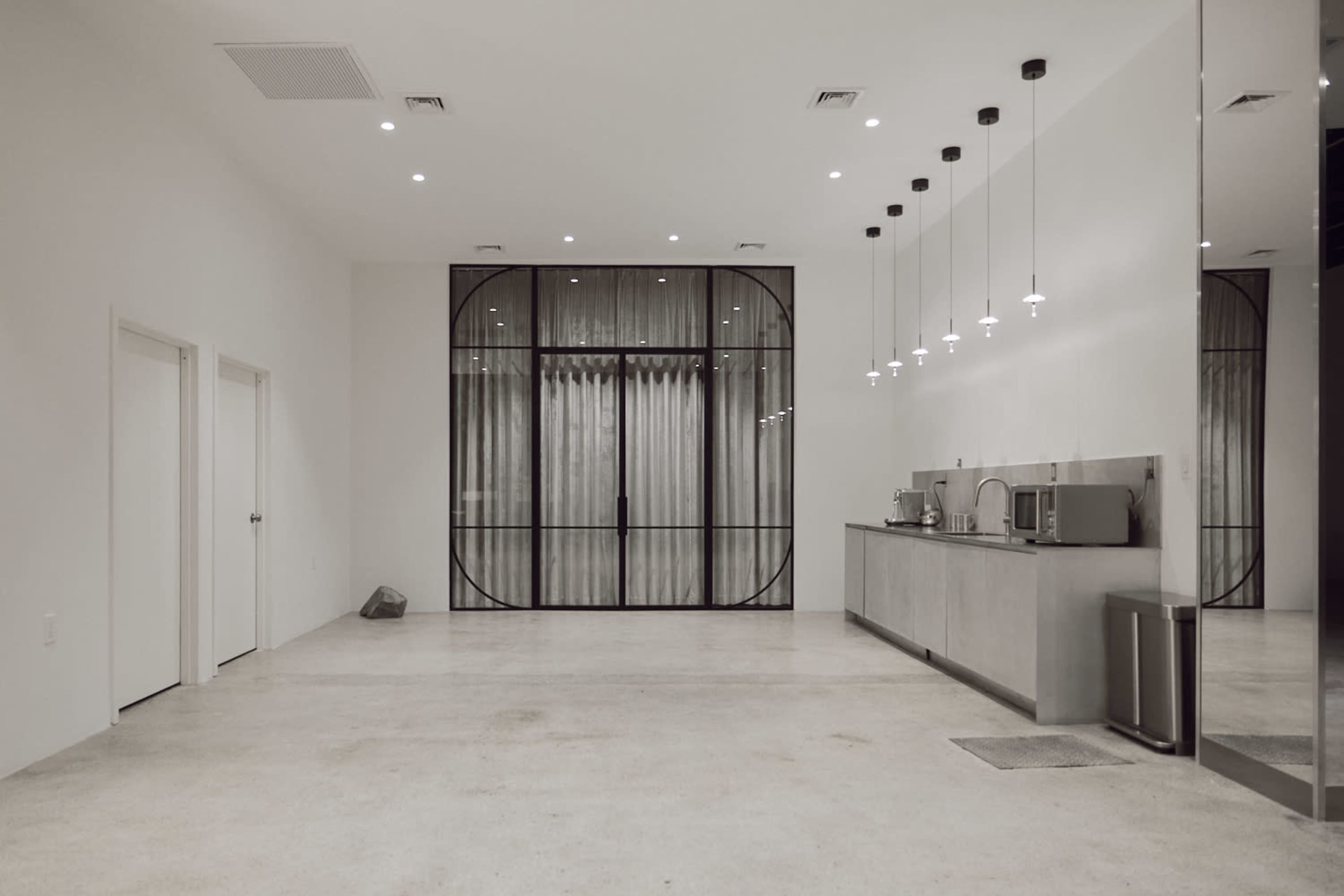 The image shows a modern, minimalist interior space featuring a brightly lit kitchen area, two closed doors, and a large glass-paneled partition with curtains at the far end.
