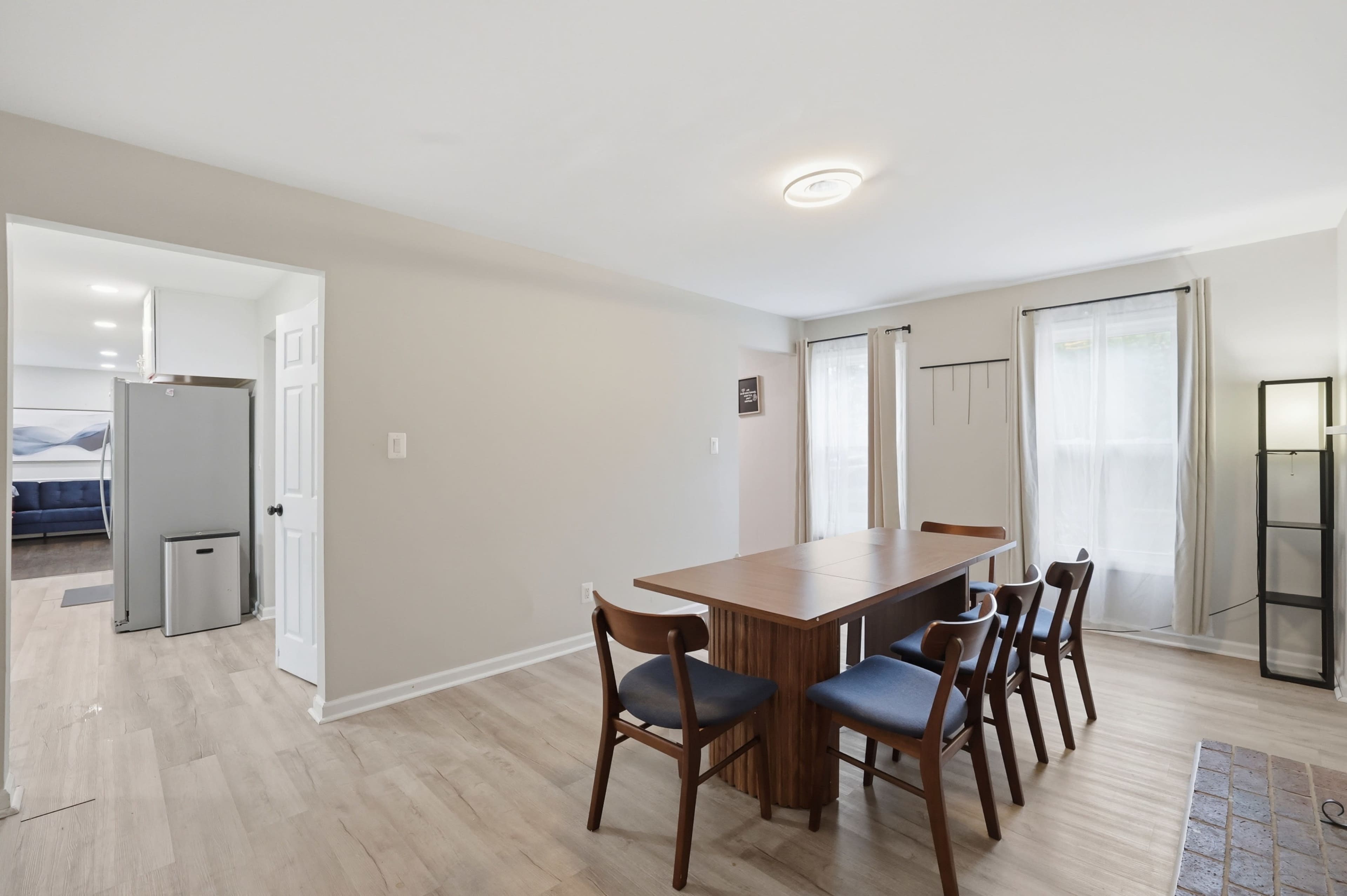A dining area features a wooden table surrounded by six chairs, with large windows letting in natural light and a doorway leading to another room.
