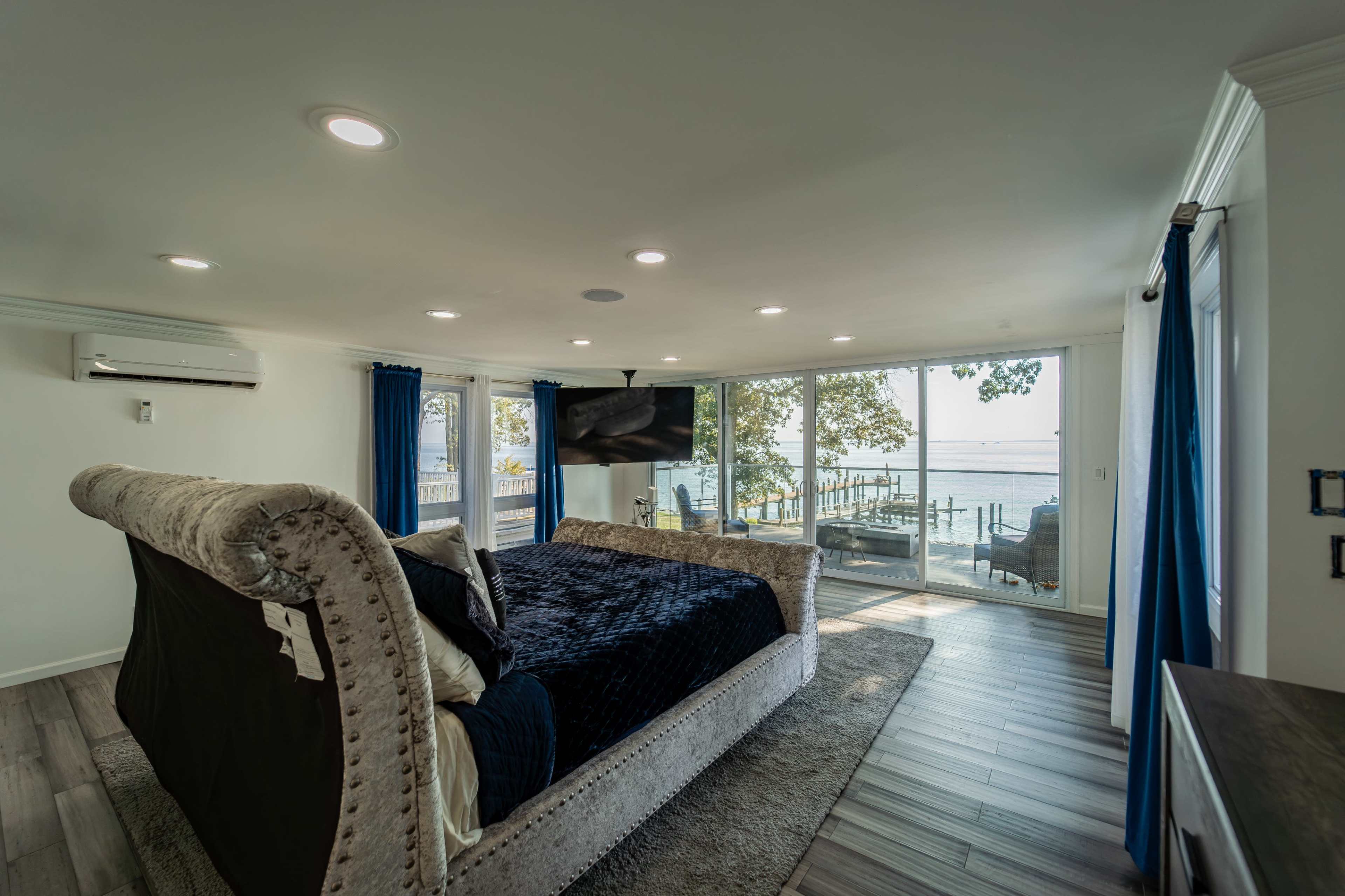 A modern bedroom with a large bed, facing sliding glass doors that open to a waterfront view.