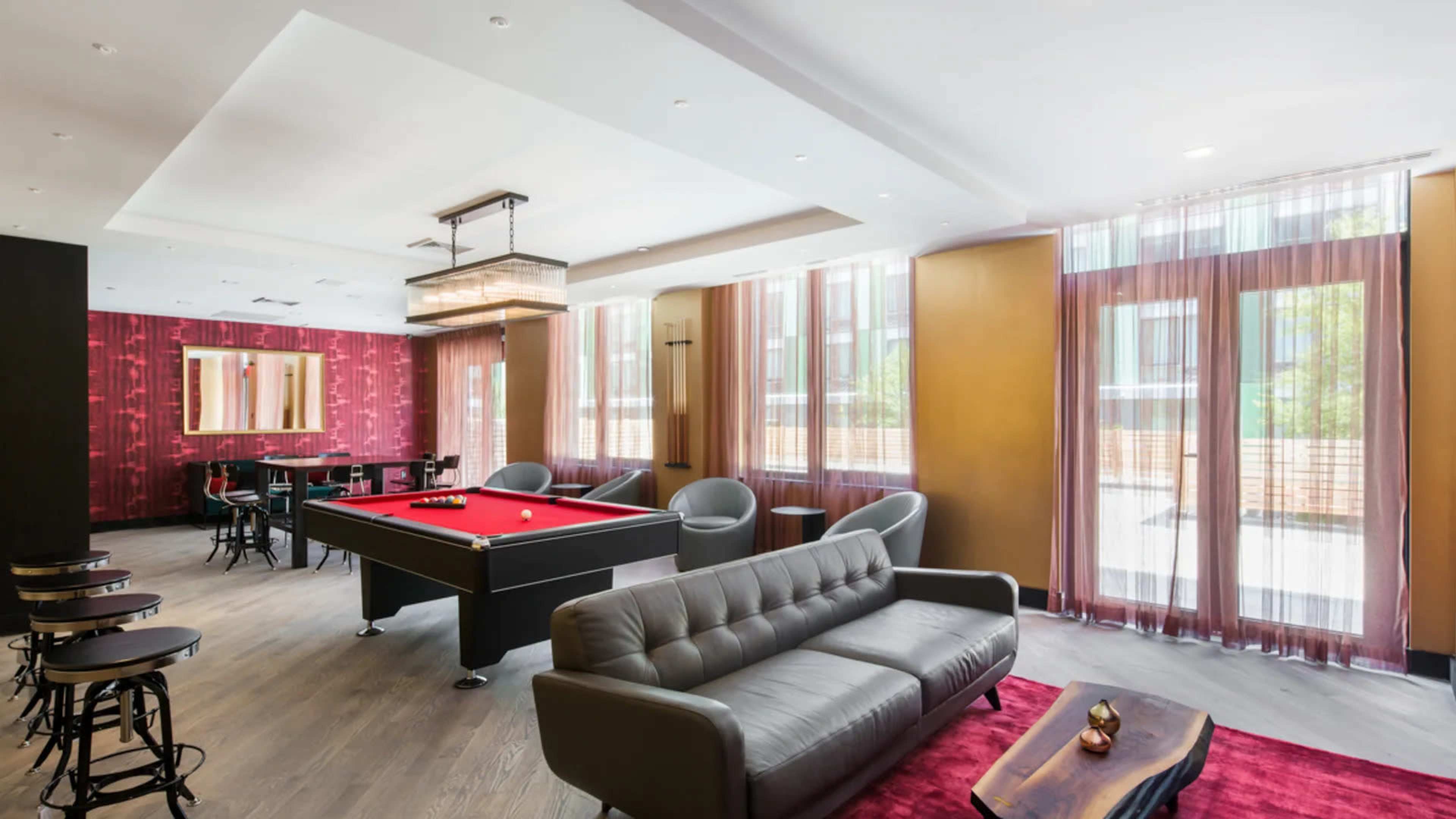 A modern lounge area featuring a billiards table, a gray couch, and large windows with sheer curtains.
