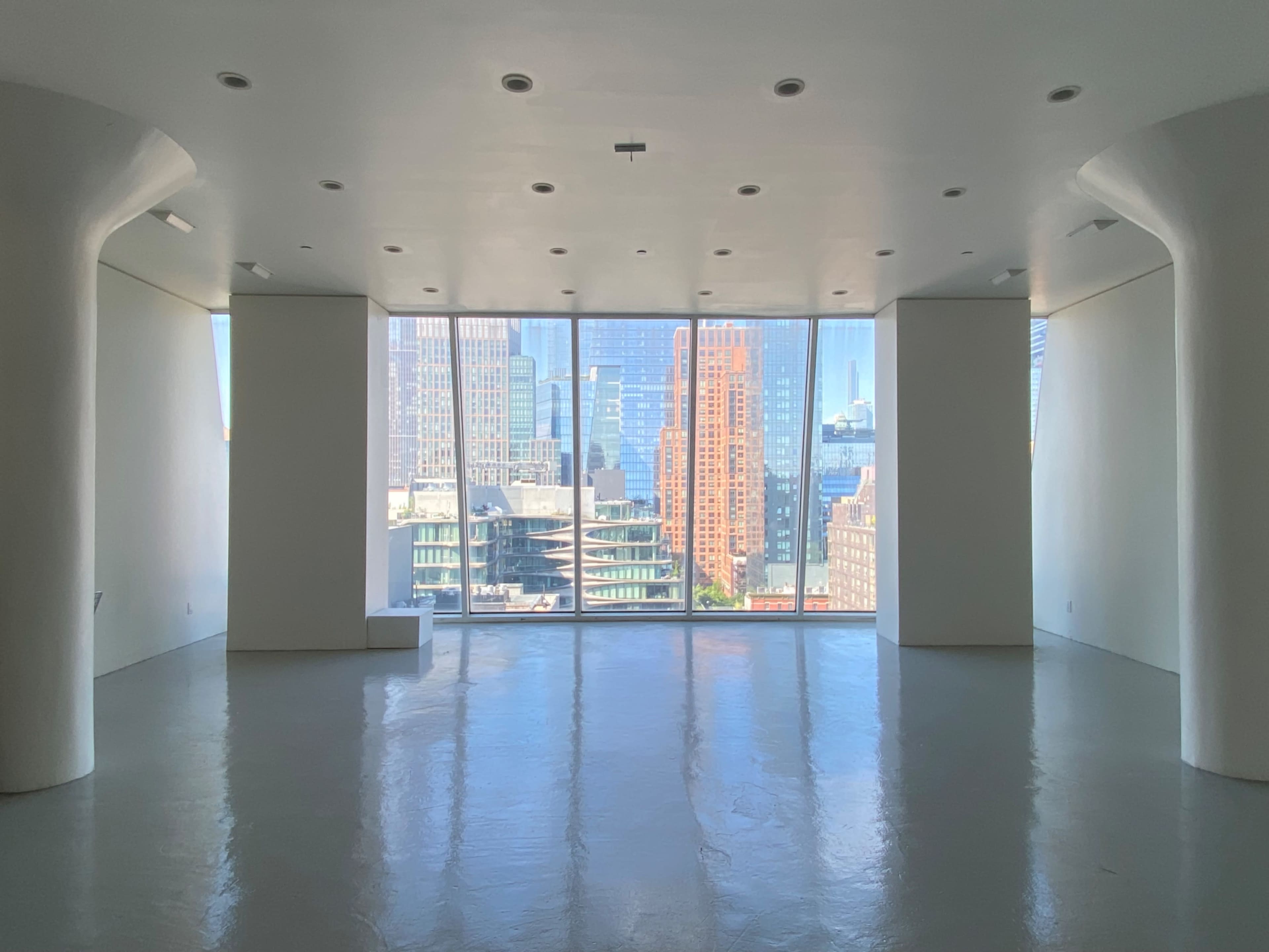 The image shows a spacious, empty modern room with large windows offering a view of urban buildings and a clear sky.