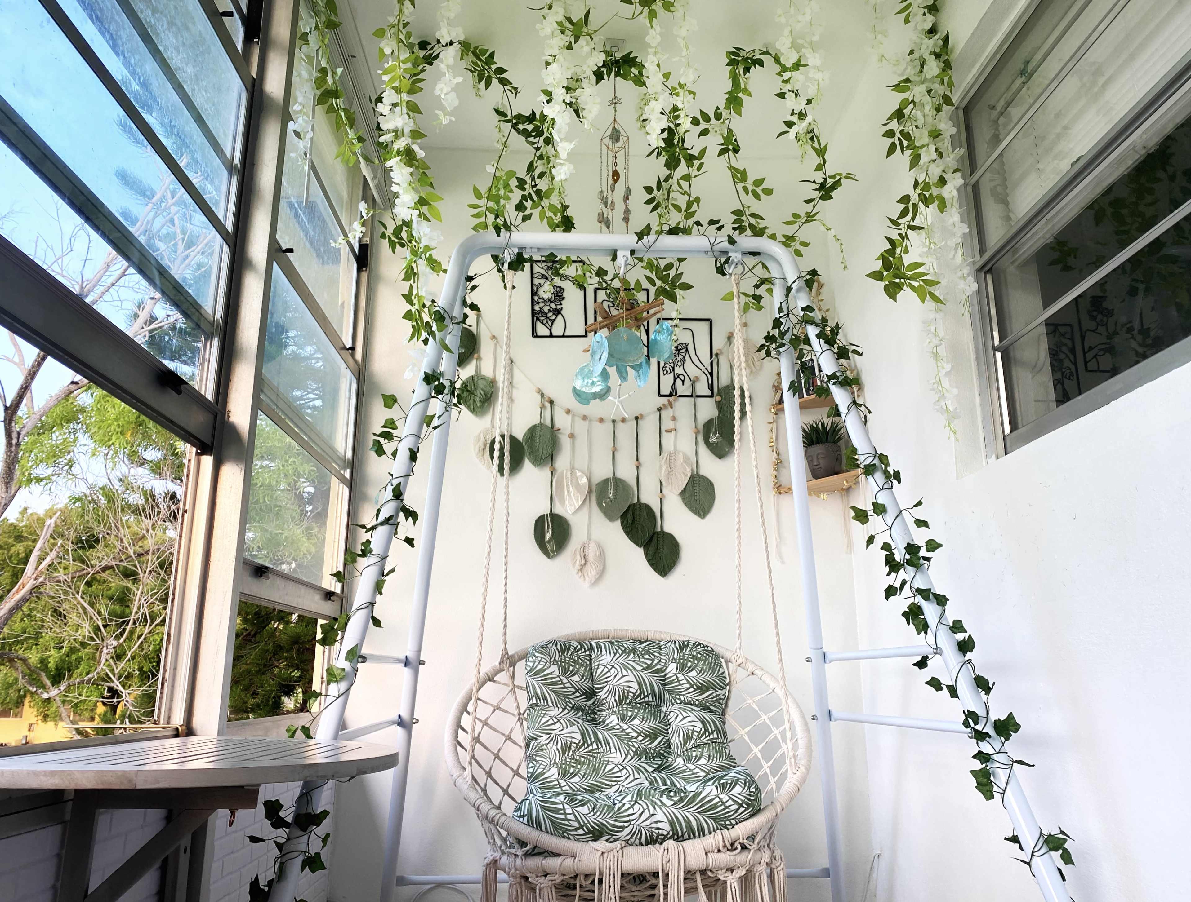A cozy balcony featuring a suspended swing chair adorned with green foliage and decorative wall hangings.
