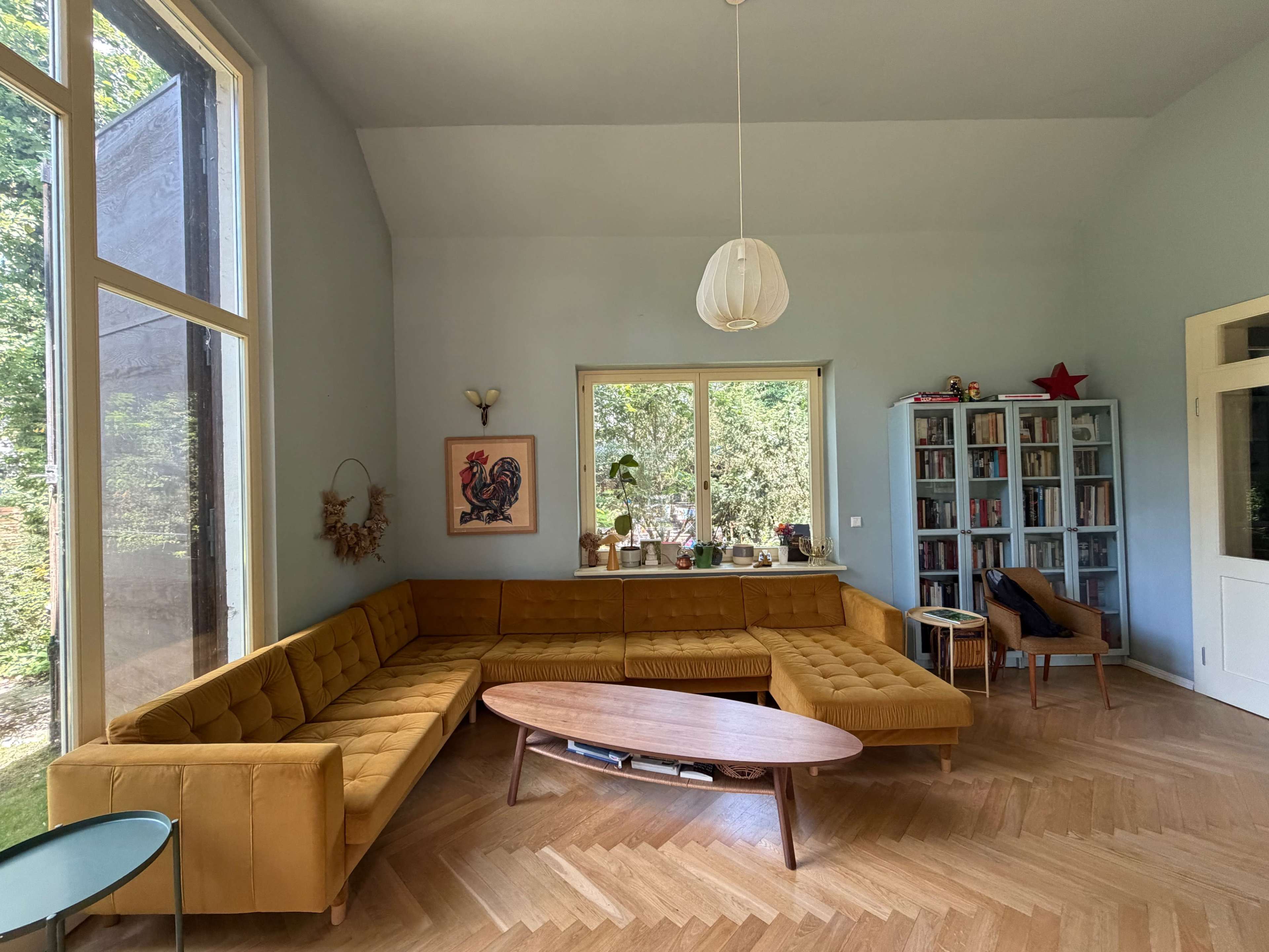 The image shows a spacious living room with a yellow sectional sofa, an oval coffee table, and a bookshelf against the wall, surrounded by large windows that let in natural light.
