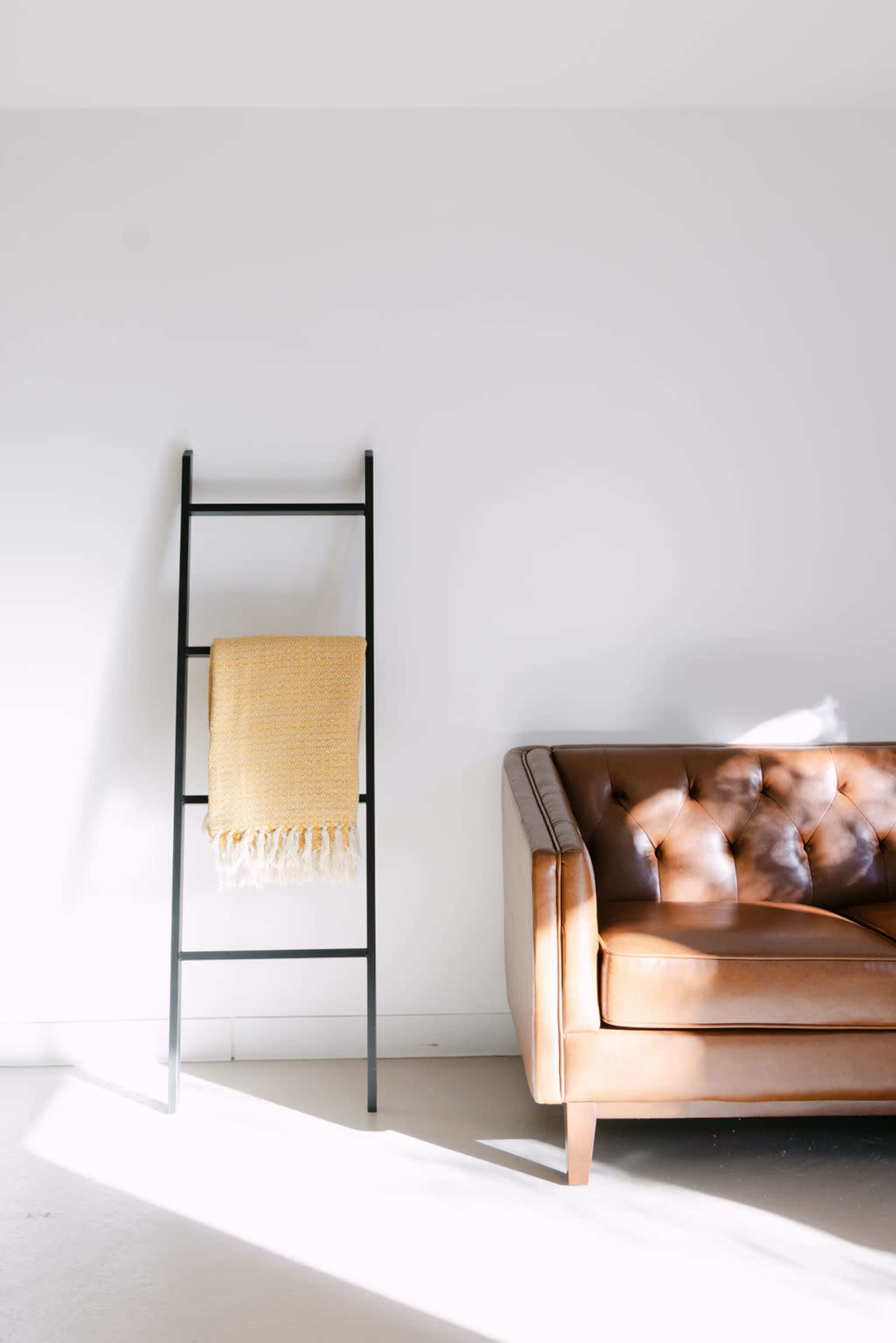 A brown leather sofa is positioned next to a black ladder with a yellow blanket draped over it against a white wall.