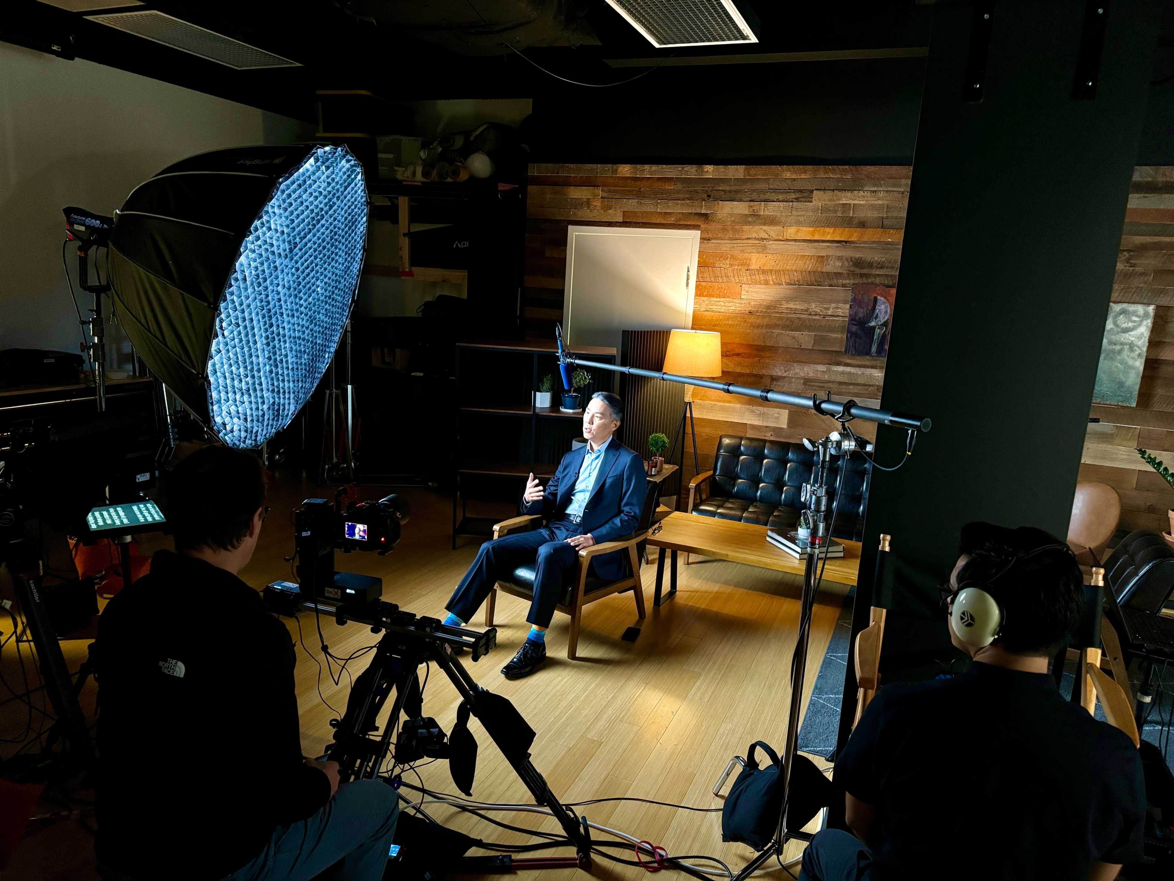A man in a suit sits on a chair in a well-lit studio, surrounded by cameras and lighting equipment while being filmed.