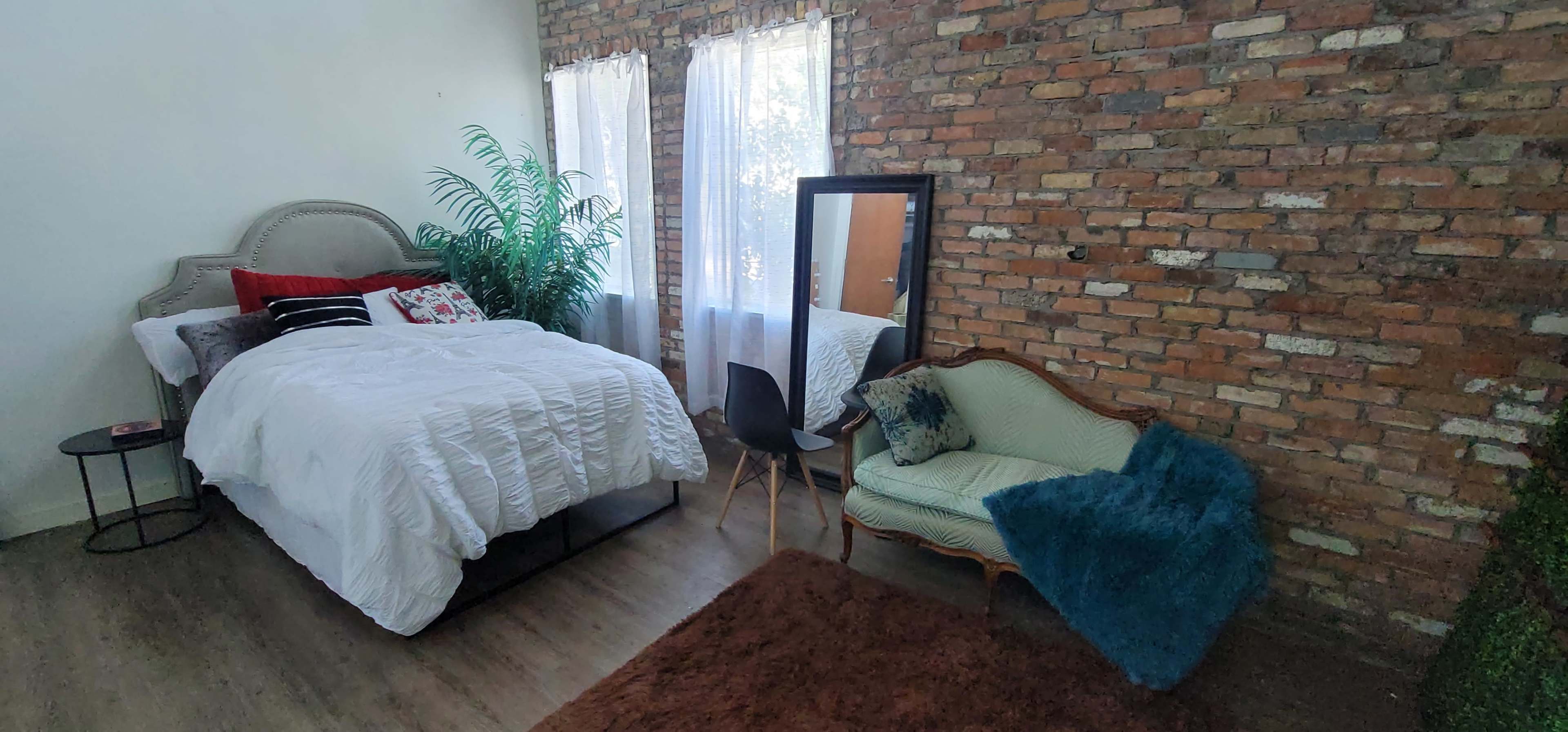 A bedroom with a brick wall, a bed with white bedding, a mirror, a green armchair, and a large potted plant near the window.