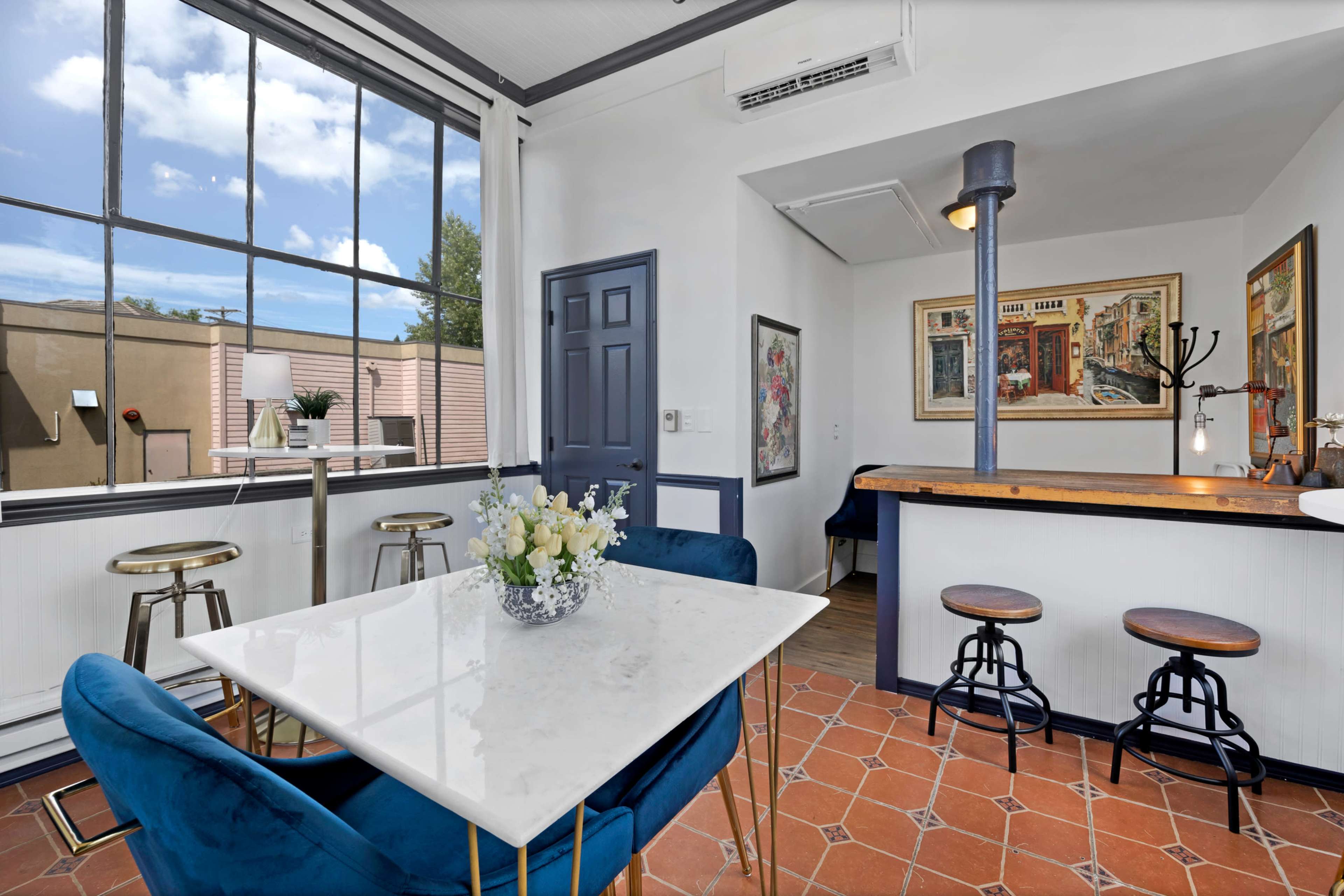 The image shows a modern indoor dining area with a marble table, blue chairs, and large windows letting in natural light, alongside a kitchen counter and bar stools.