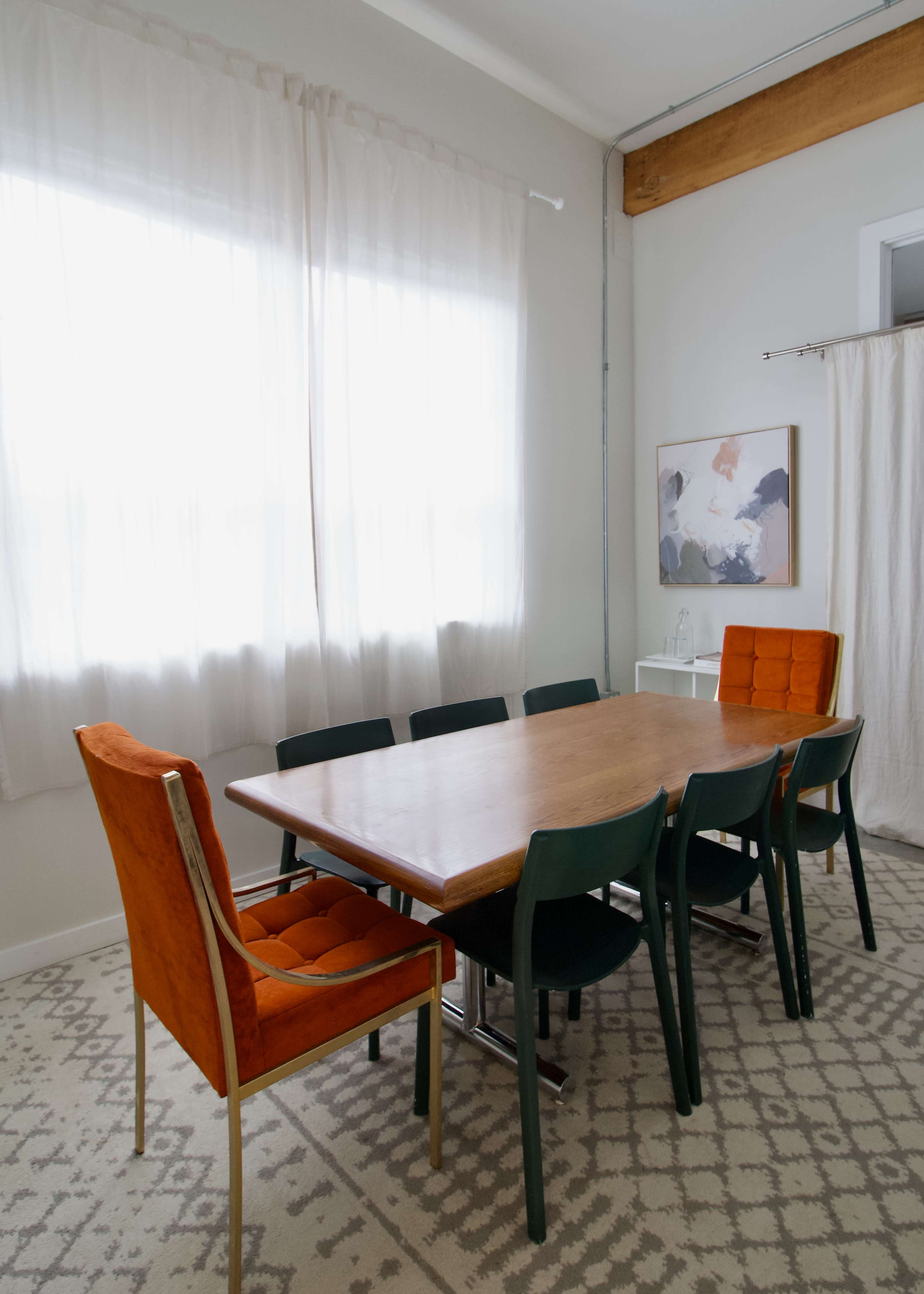 A dining room features a wooden table surrounded by six chairs, with large windows draped in sheer curtains and a piece of artwork on the wall.