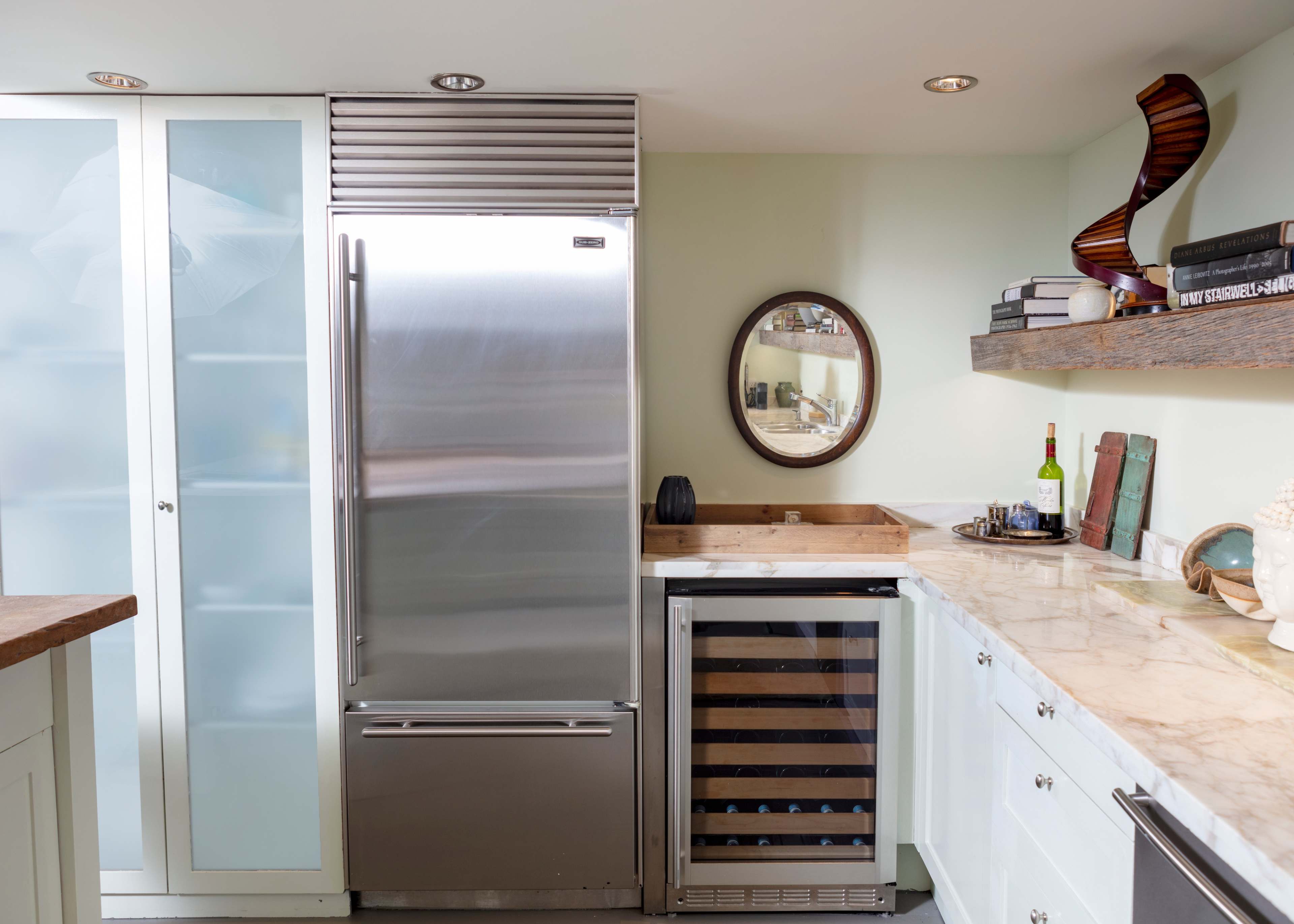A modern kitchen featuring stainless steel appliances, including a refrigerator and a wine cooler, with a marble countertop and glass-front cabinets.