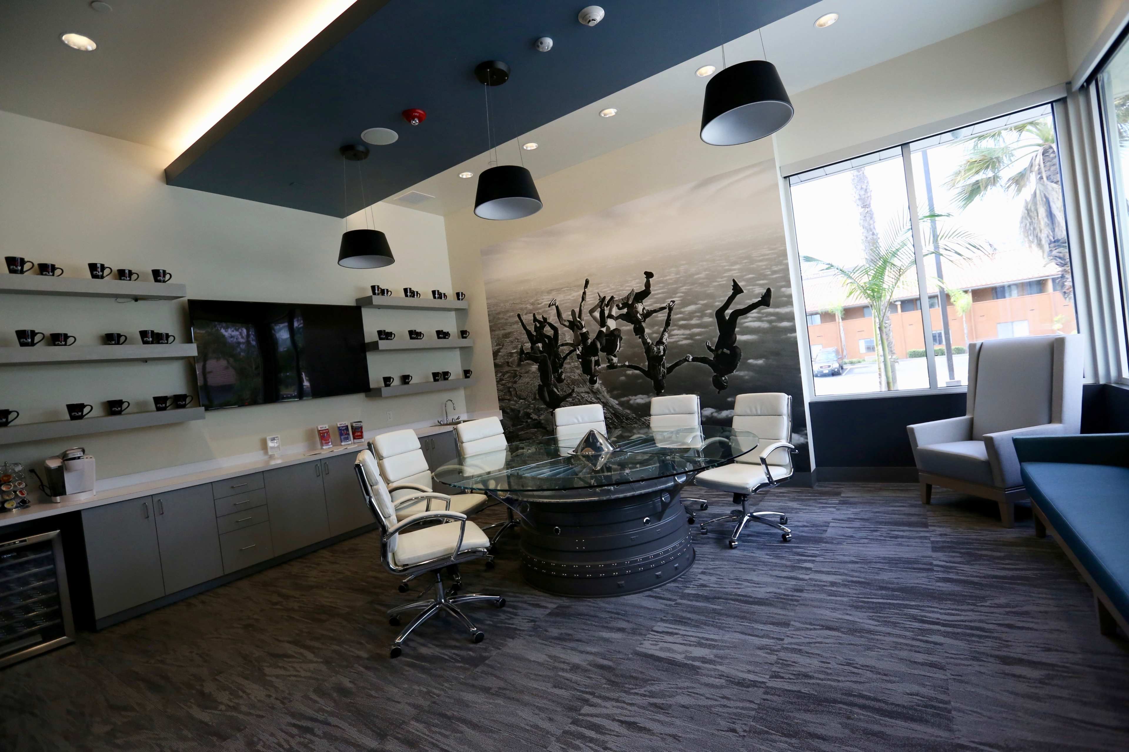 A modern conference room with a round glass table surrounded by white chairs, a wall decorated with a black-and-white mural, and a kitchenette area along one side.