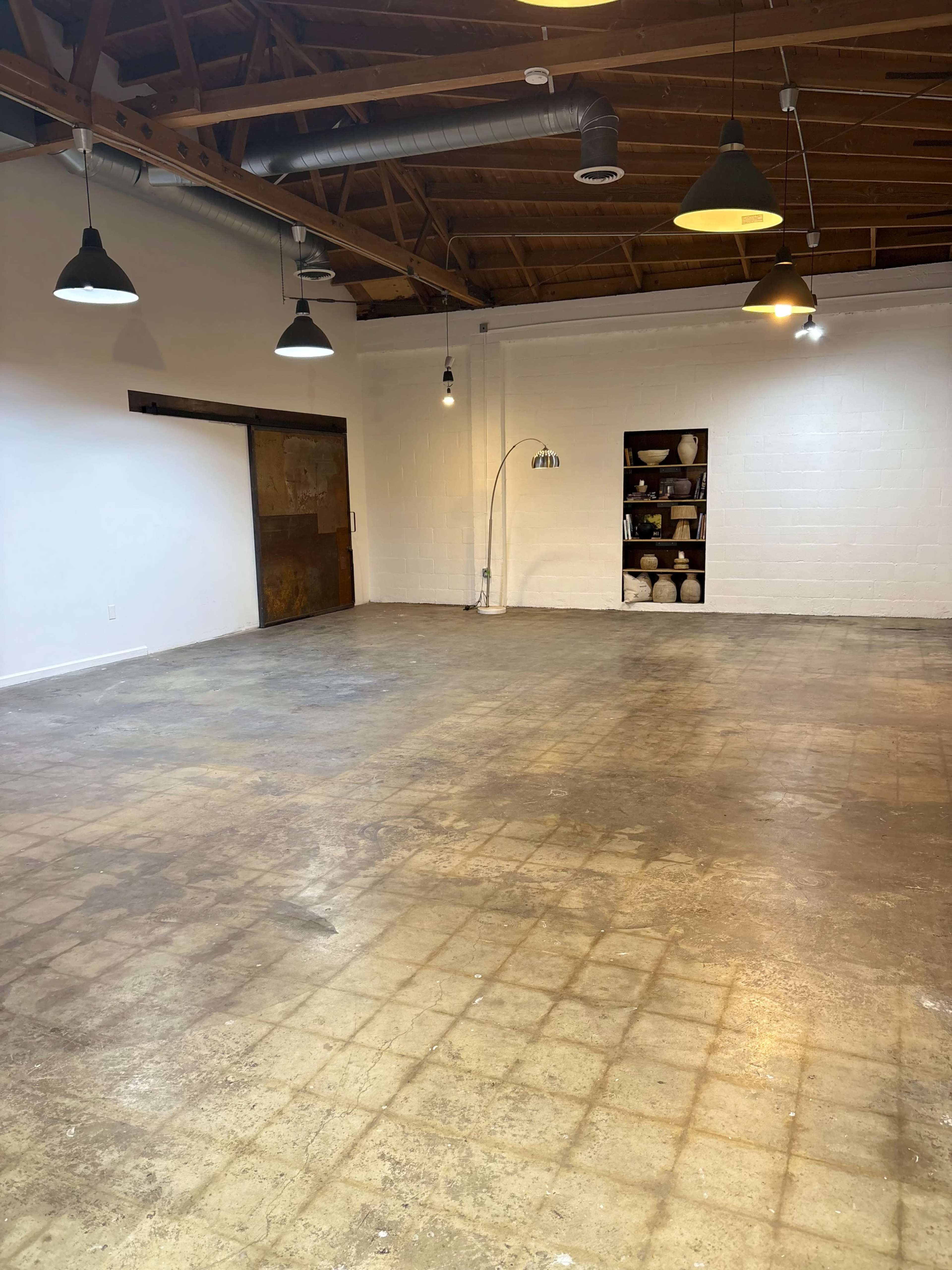 The image shows an empty, spacious room with exposed wooden beams, concrete flooring, and several hanging lights, along with a shelf displaying pottery in the corner.