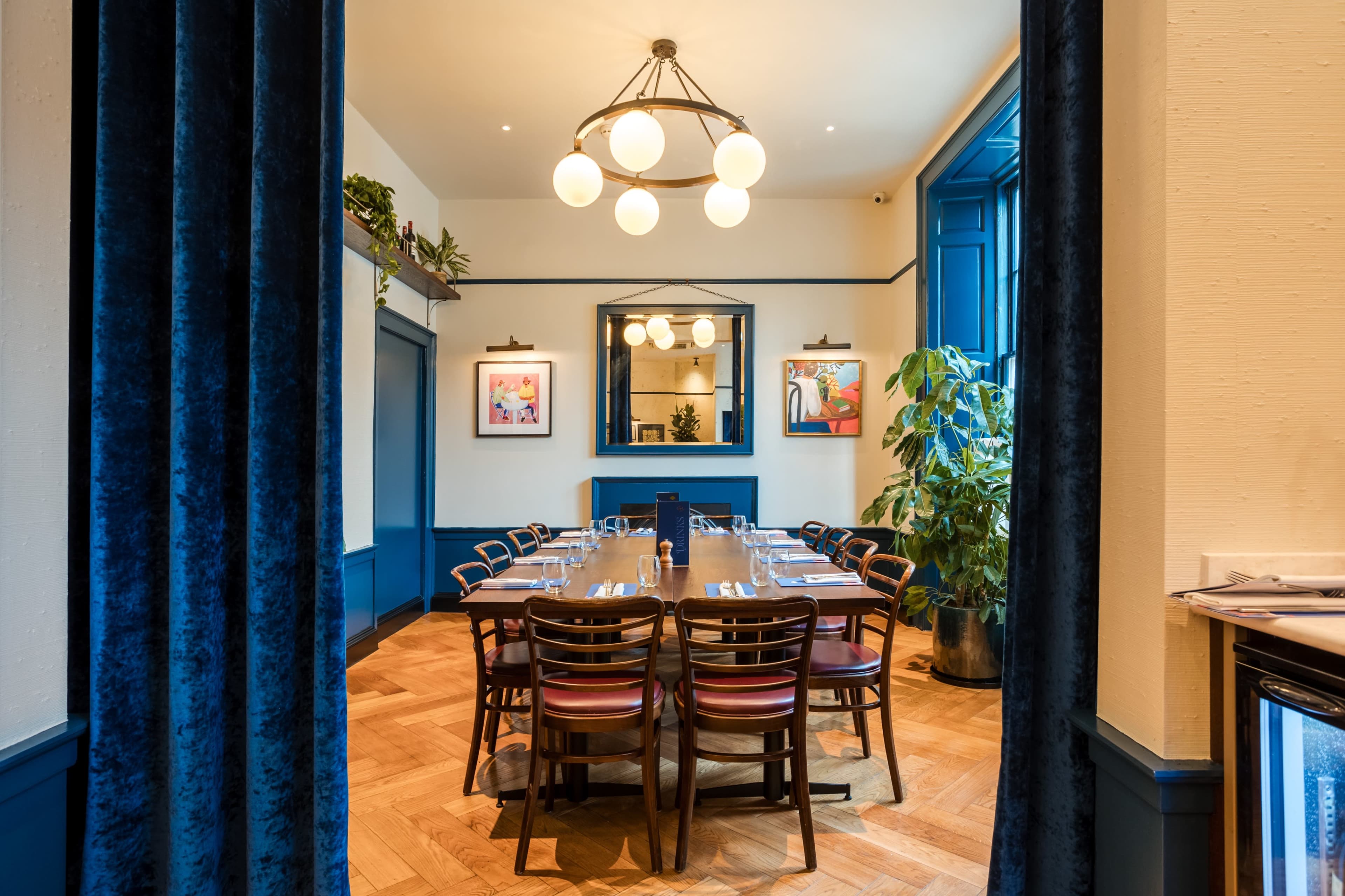 The image shows a warmly lit dining room featuring a large wooden table surrounded by multiple chairs, with decorative artwork on the walls and lush green plants in the corner.