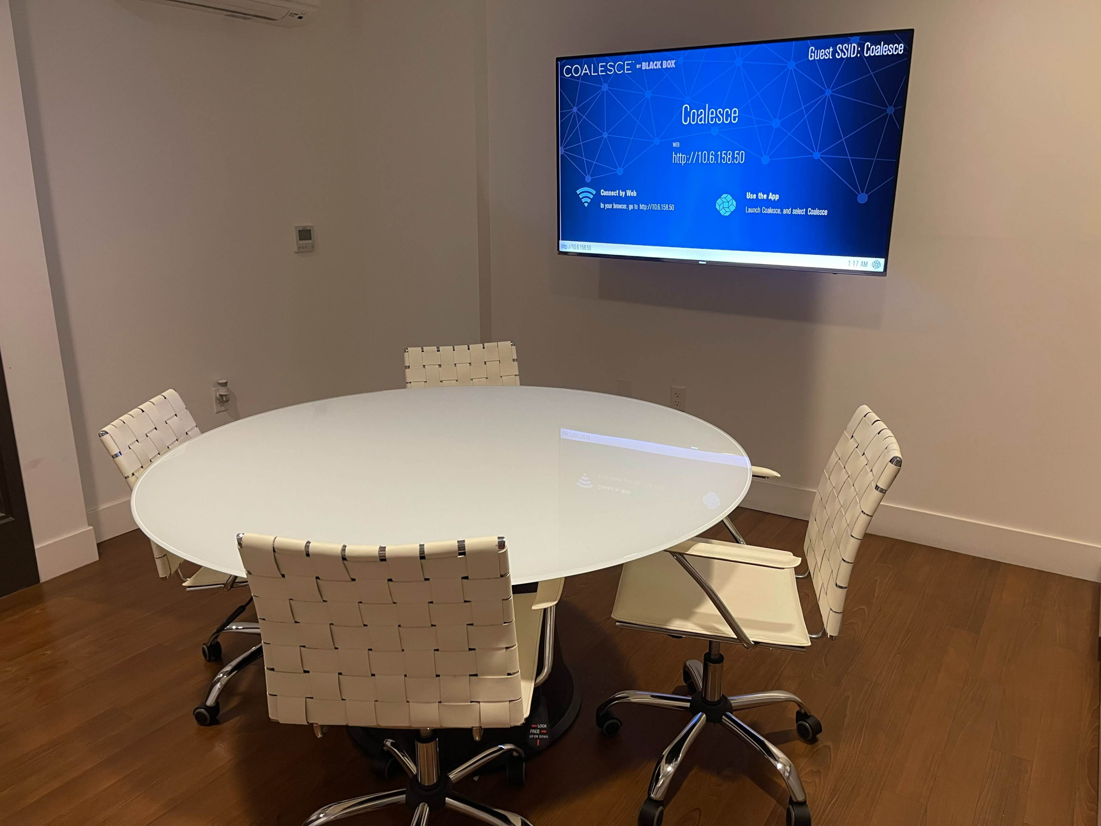 A modern conference room features a round glass table surrounded by four white chairs, with a large screen displaying information on the wall.