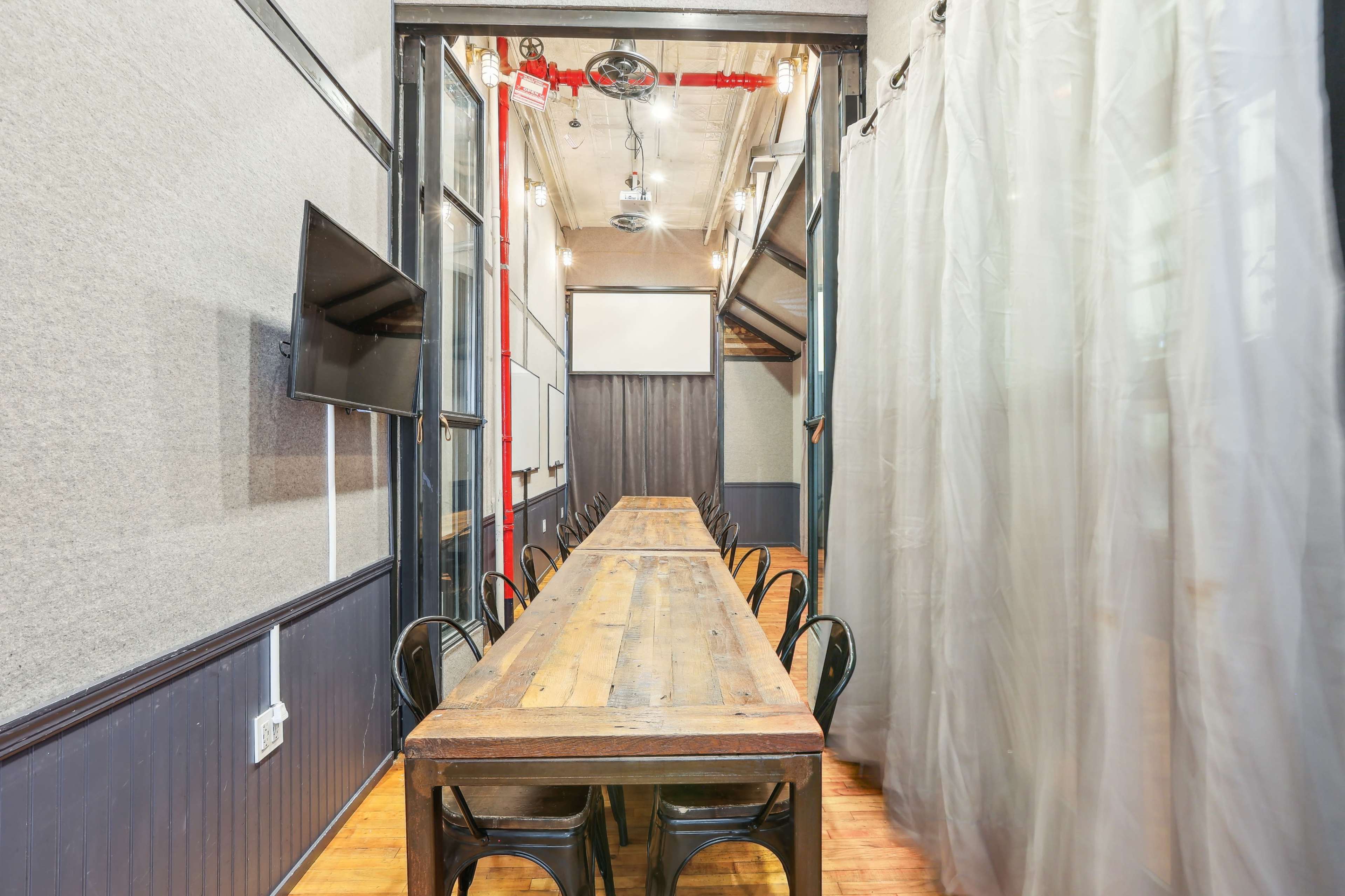 The image shows a long wooden table set in a narrow meeting room with gray walls, black chairs, and a large screen on one wall.