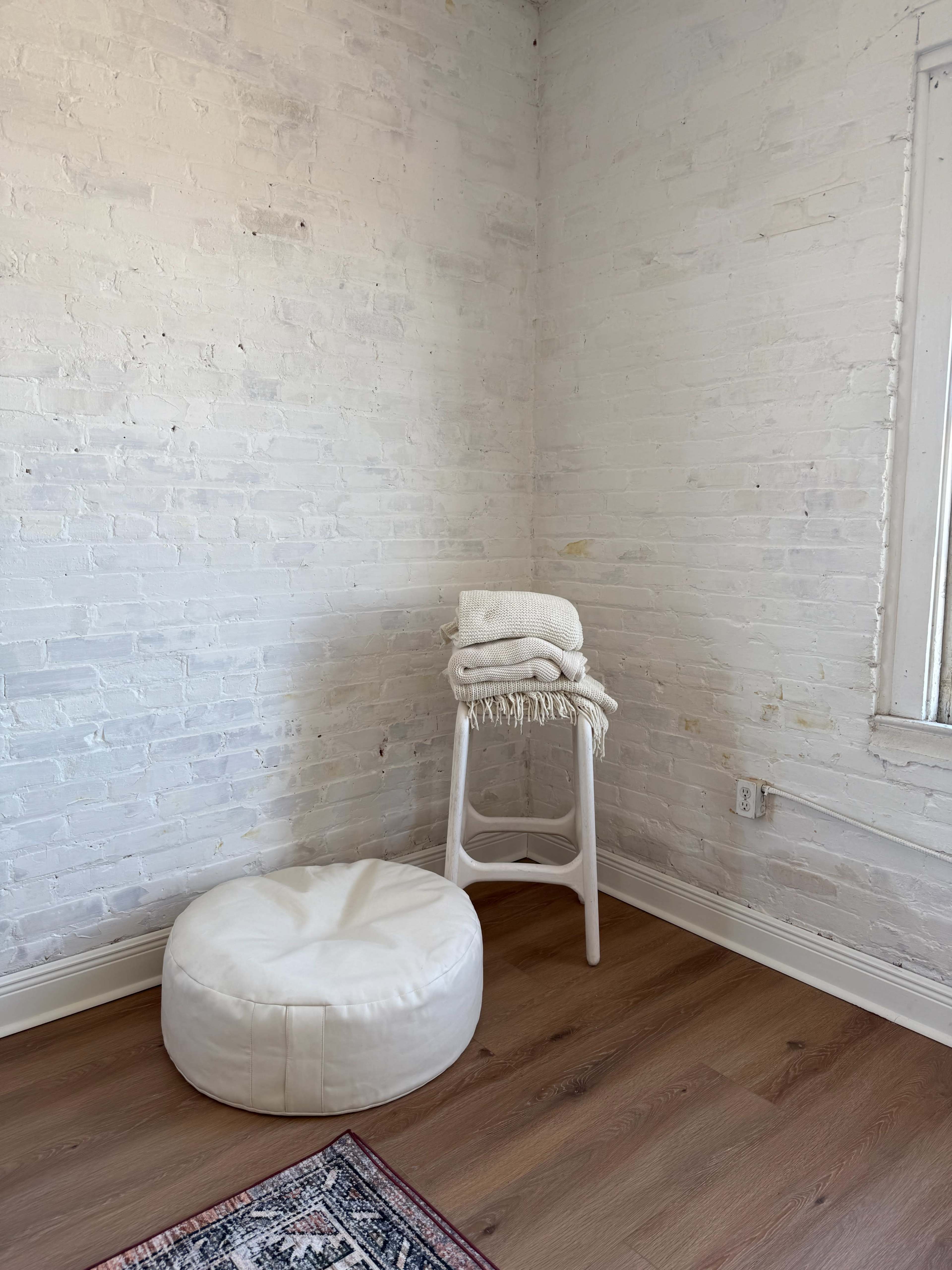 The image shows a corner of a room with a white brick wall, a stacked towel on a stool, and a white pouf on the floor.