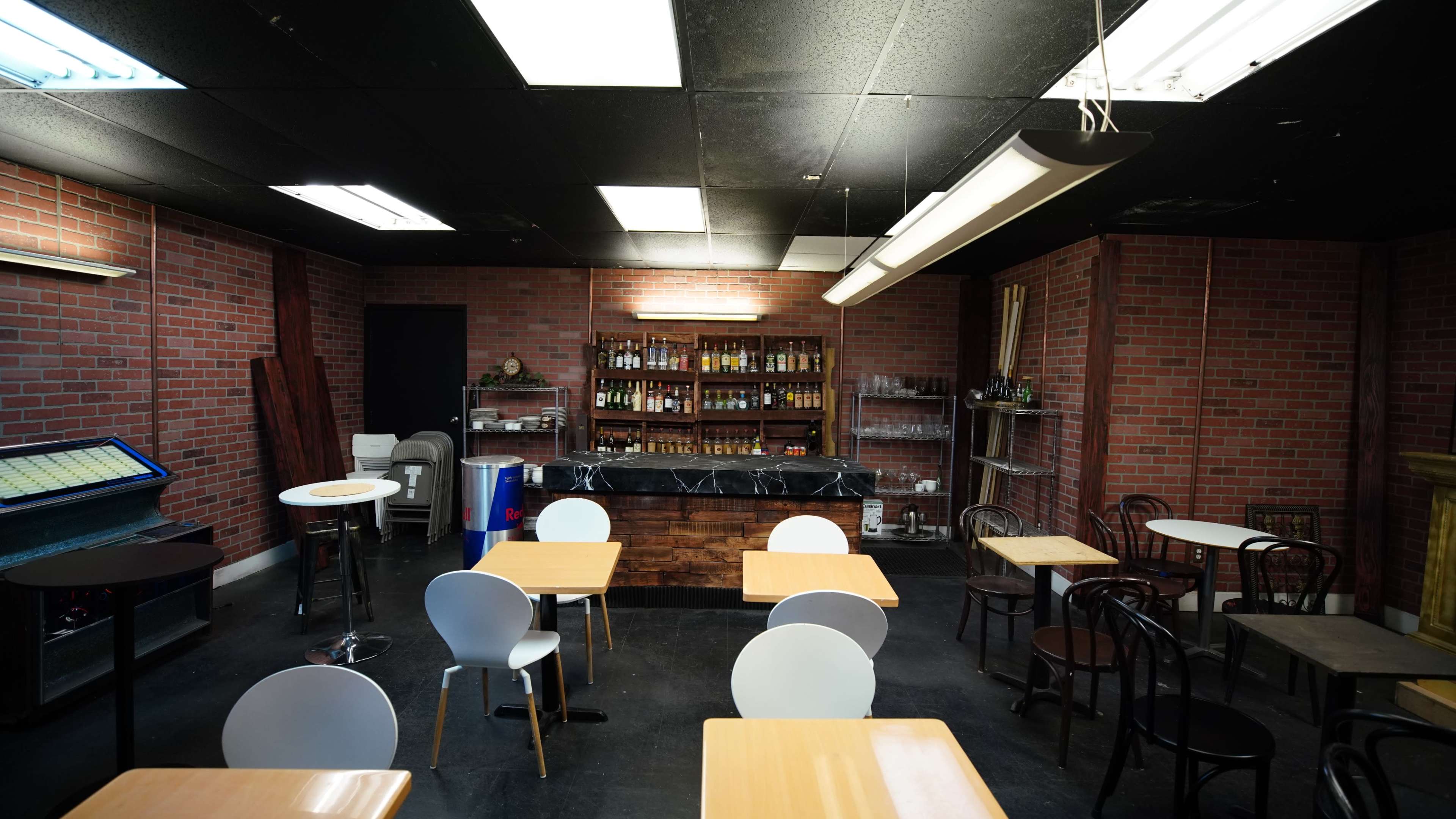 A cafe interior with brick walls, a bar counter stocked with bottles, and wooden tables and chairs arranged around the space.