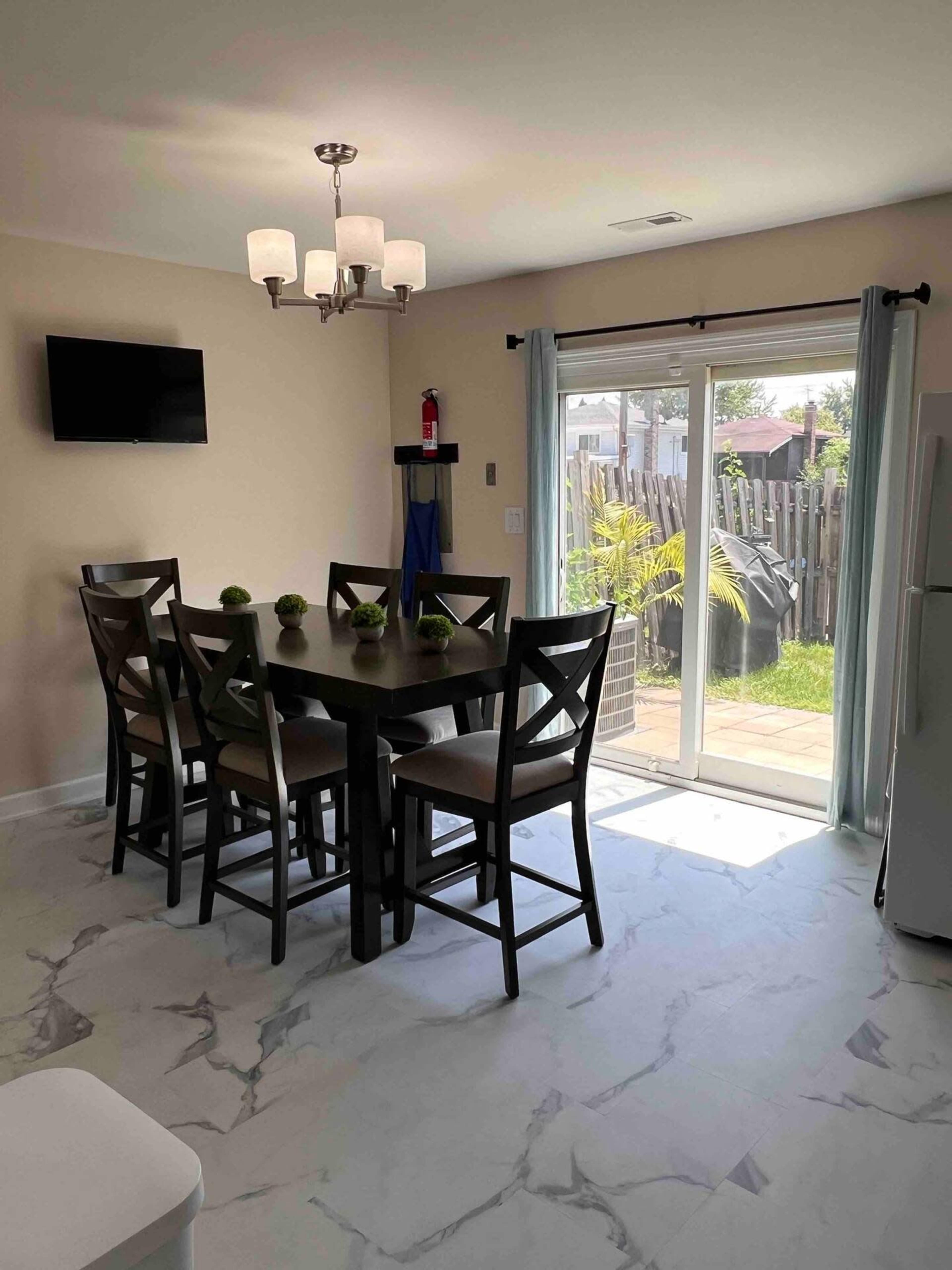A dining area features a rectangular table with six chairs, a chandelier above, and a sliding glass door leading to an outdoor space.