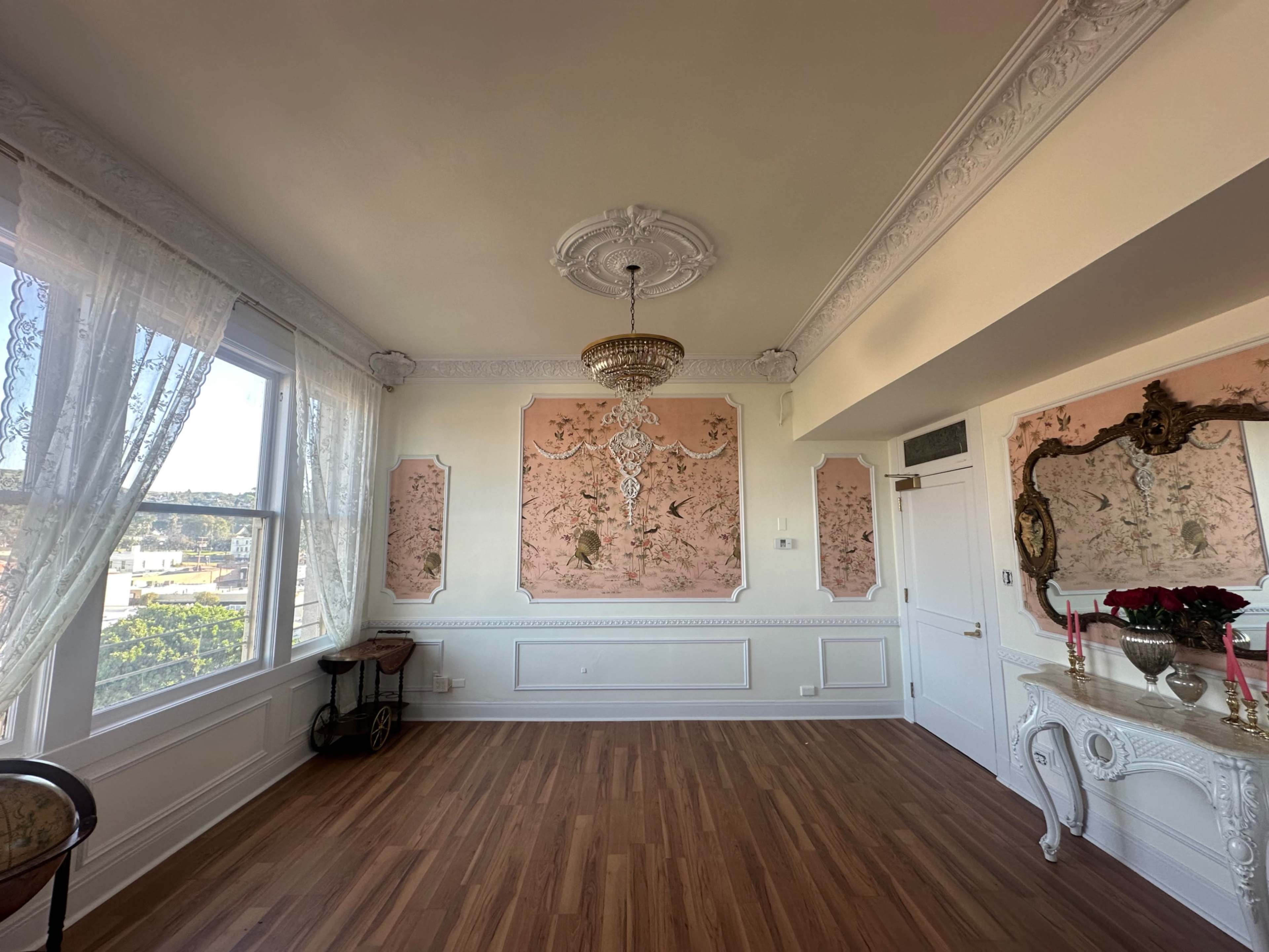 The room features ornate wall decorations, a chandelier, and large windows with sheer curtains, all set against a backdrop of polished wooden flooring.