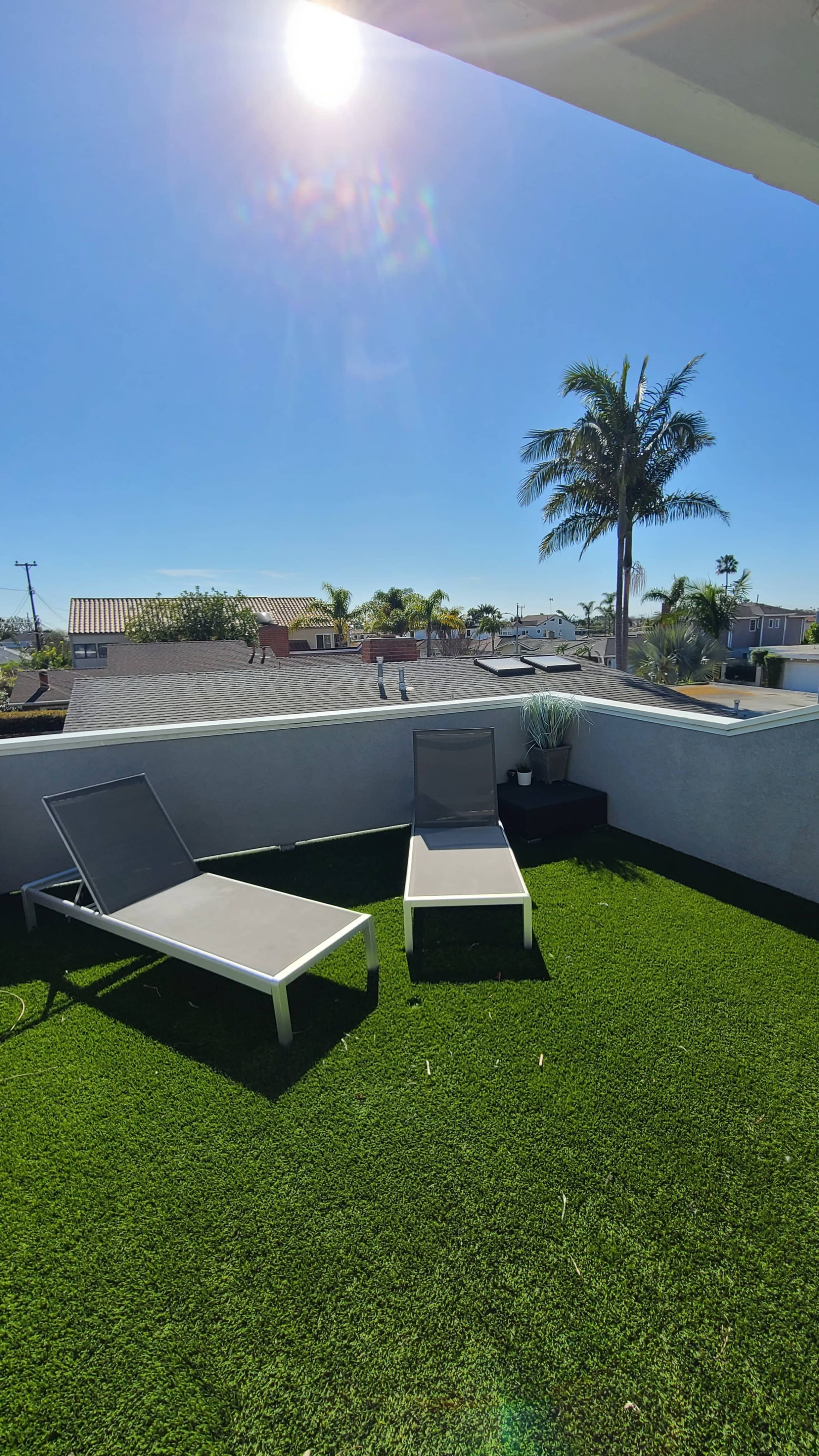 Two lounge chairs on a terrace with artificial grass, overlooking a neighborhood and palm trees under a clear blue sky.