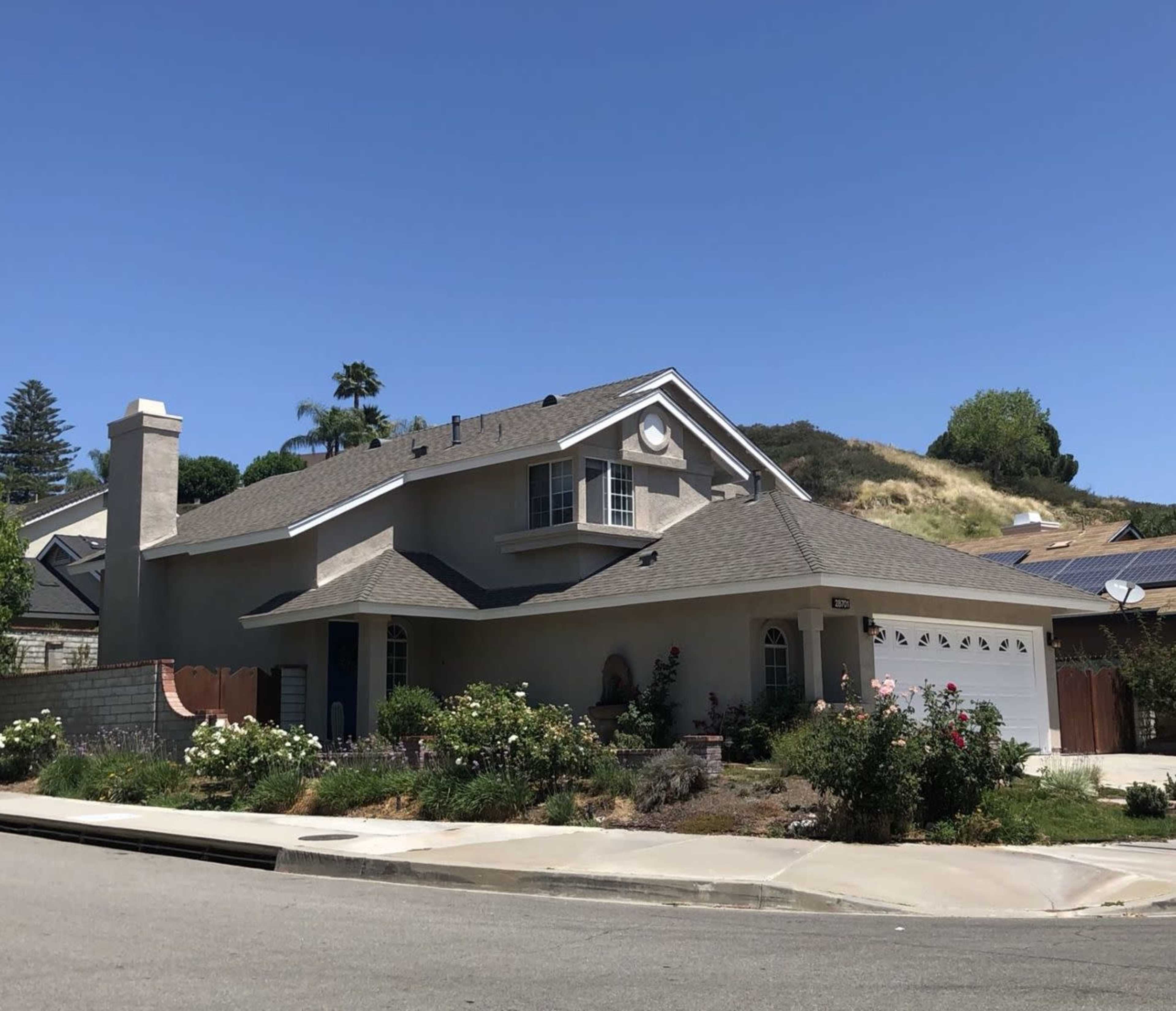 A two-story house with a gray exterior, white trim, and a landscaped front yard sits on a corner lot under a clear blue sky.
