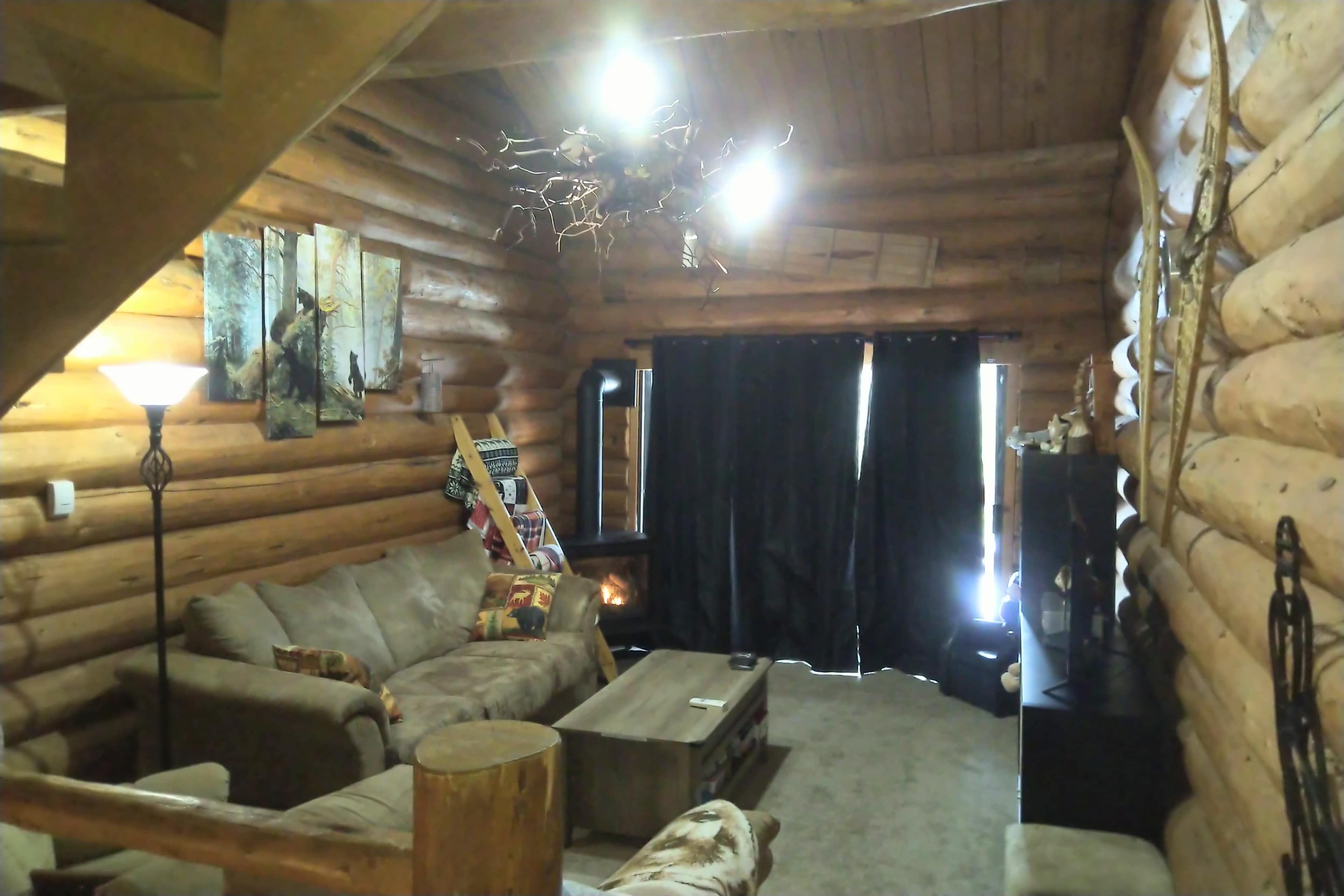 The interior of a log cabin features a cozy living area with a sofa, wooden coffee table, and a staircase leading to a loft, illuminated by several light fixtures.