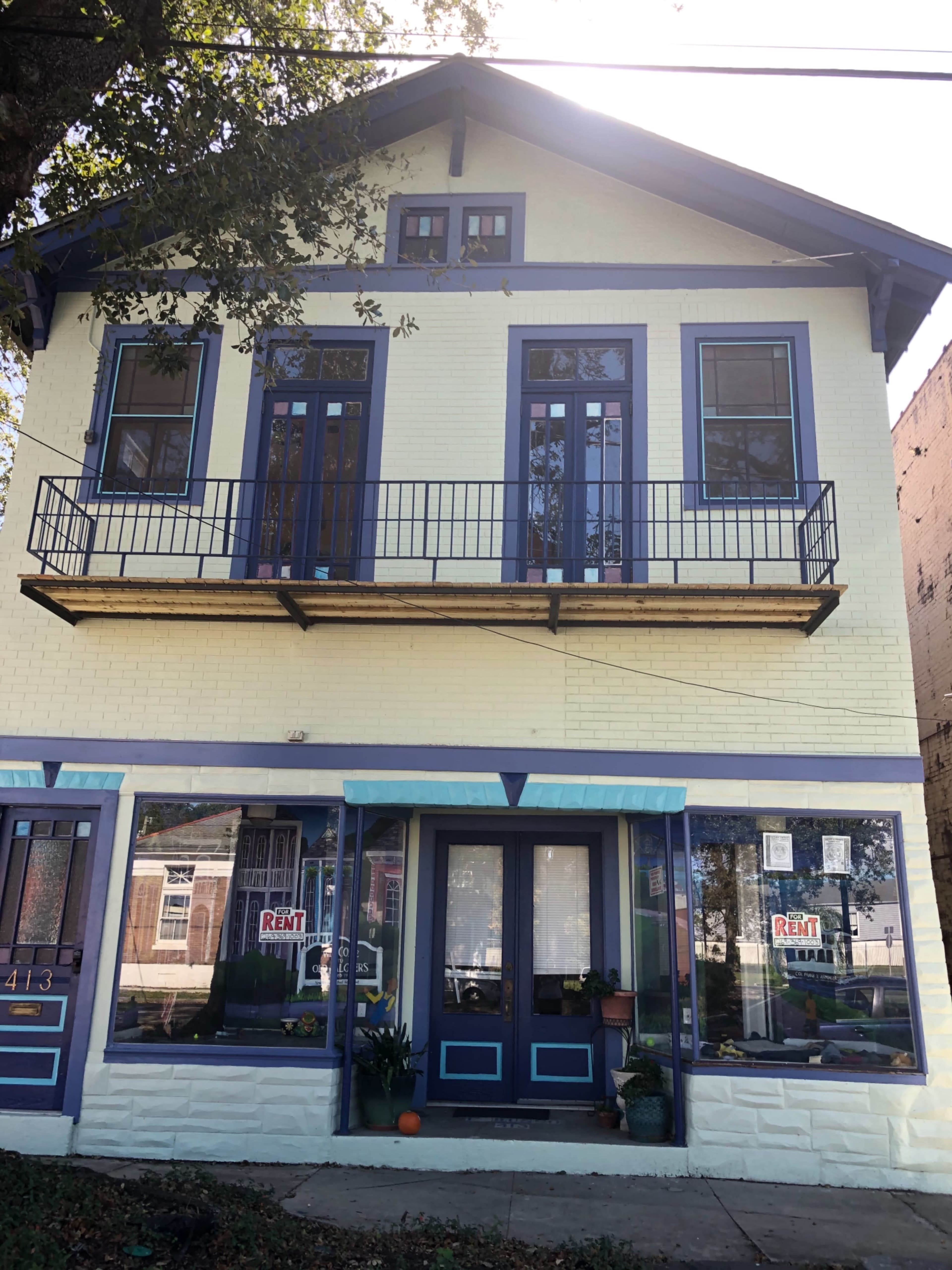 A two-story, yellow building with blue trim, featuring large windows and a "For Rent" sign in the front.