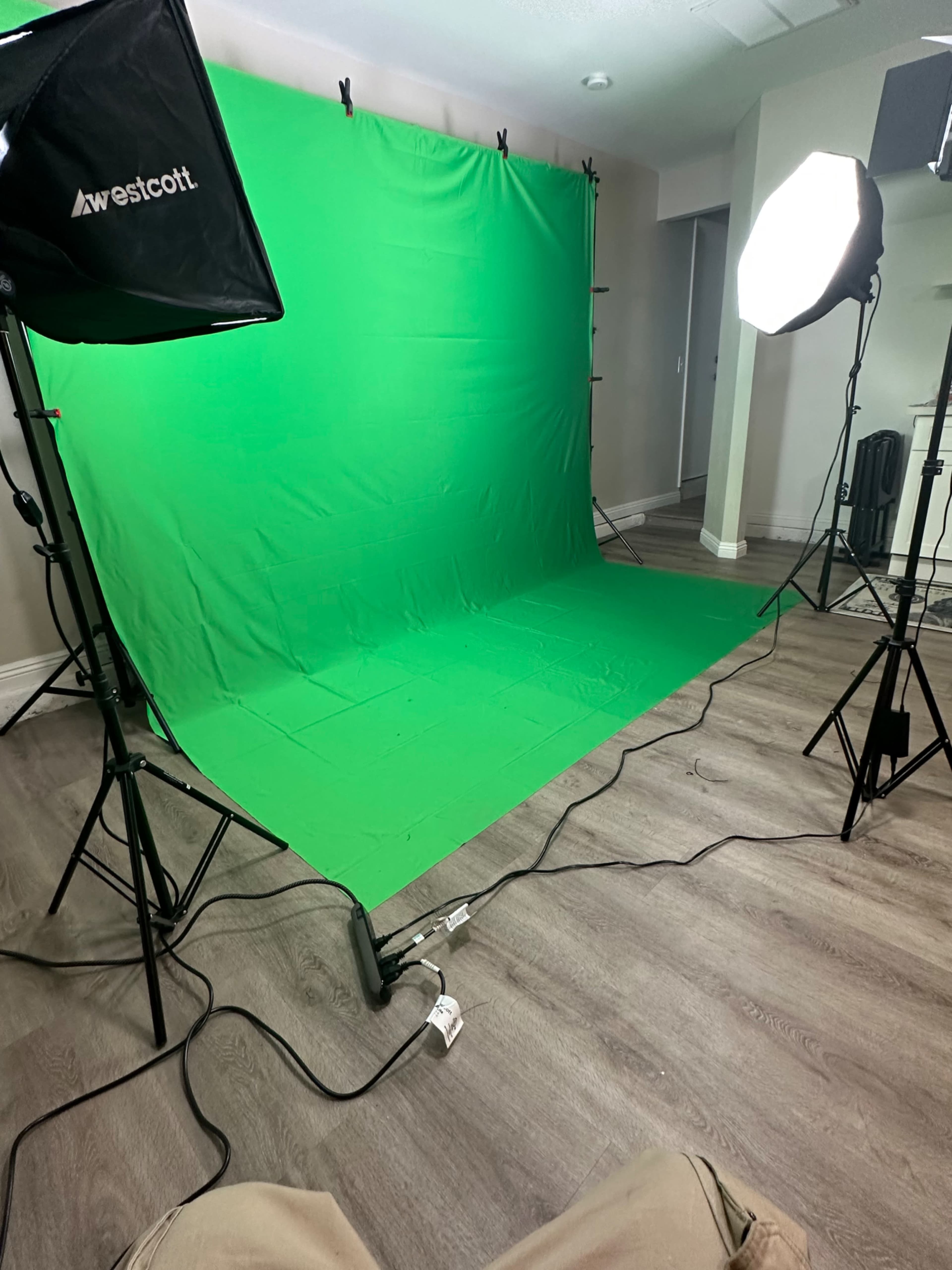 A green screen setup with softbox lights on either side, placed on a wooden floor in a well-lit room.