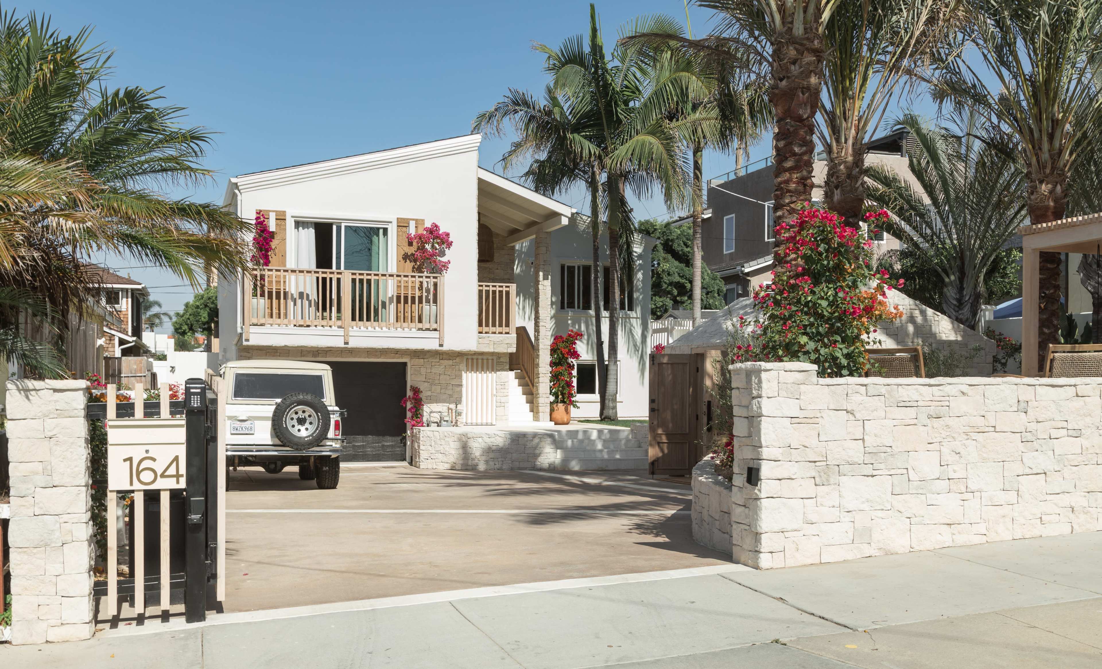 A modern two-story house with a front balcony is surrounded by palm trees and vibrant flowers in a residential area.