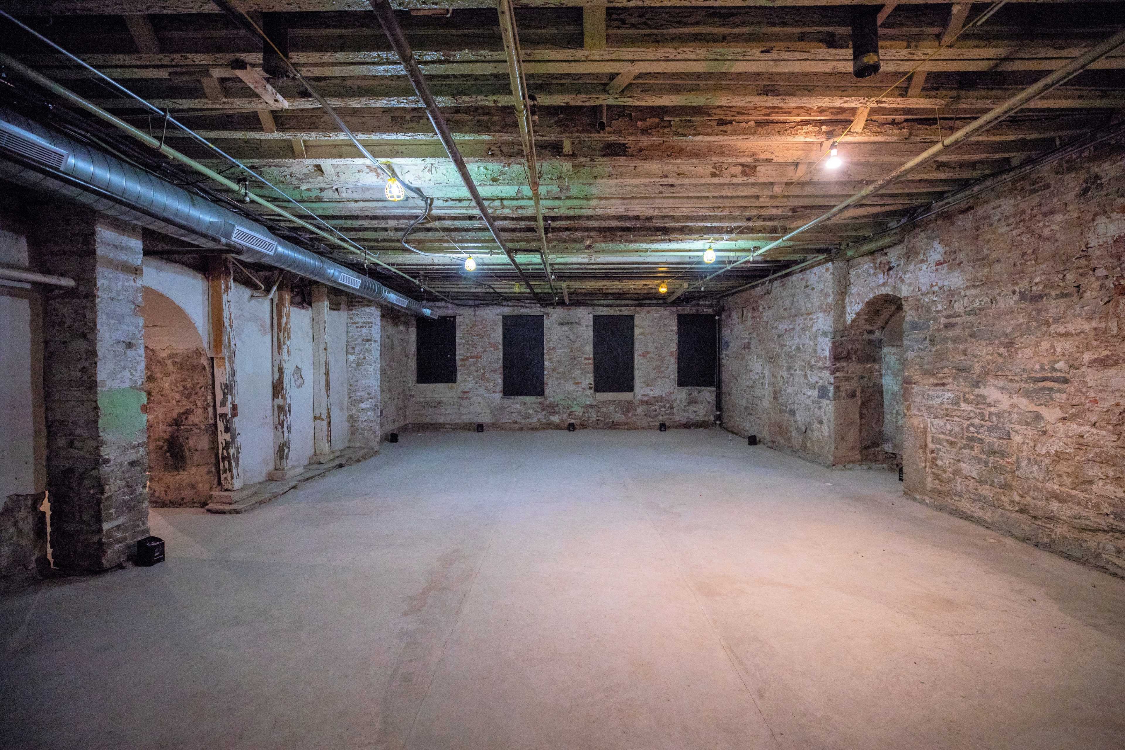 The image shows a large, empty basement room with exposed stone walls, a concrete floor, and overhead lighting.