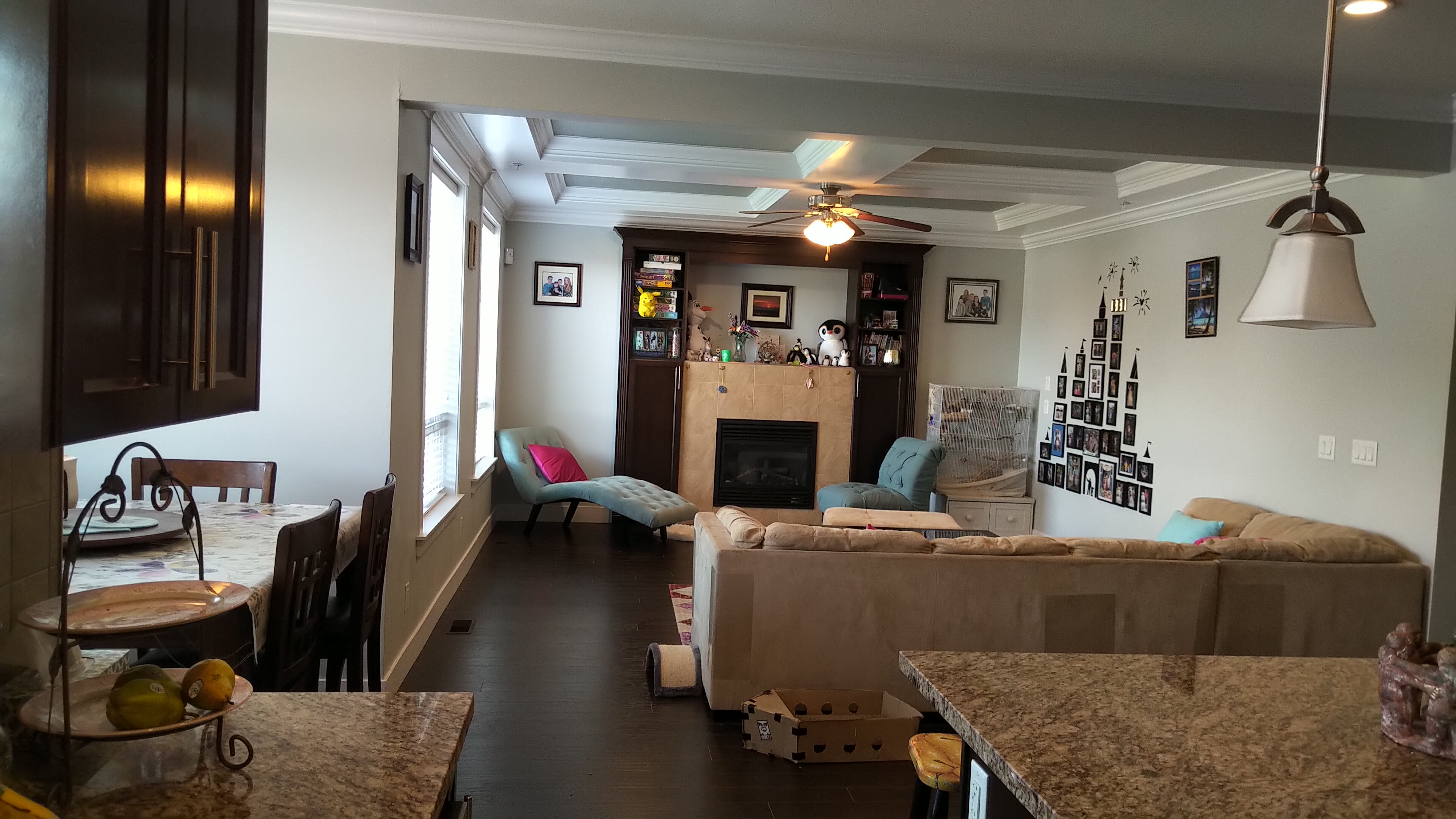A modern living room with a cream-colored sofa, a fireplace, and various decorations on the walls, while a kitchen area with dark cabinets and a dining table is visible in the foreground.