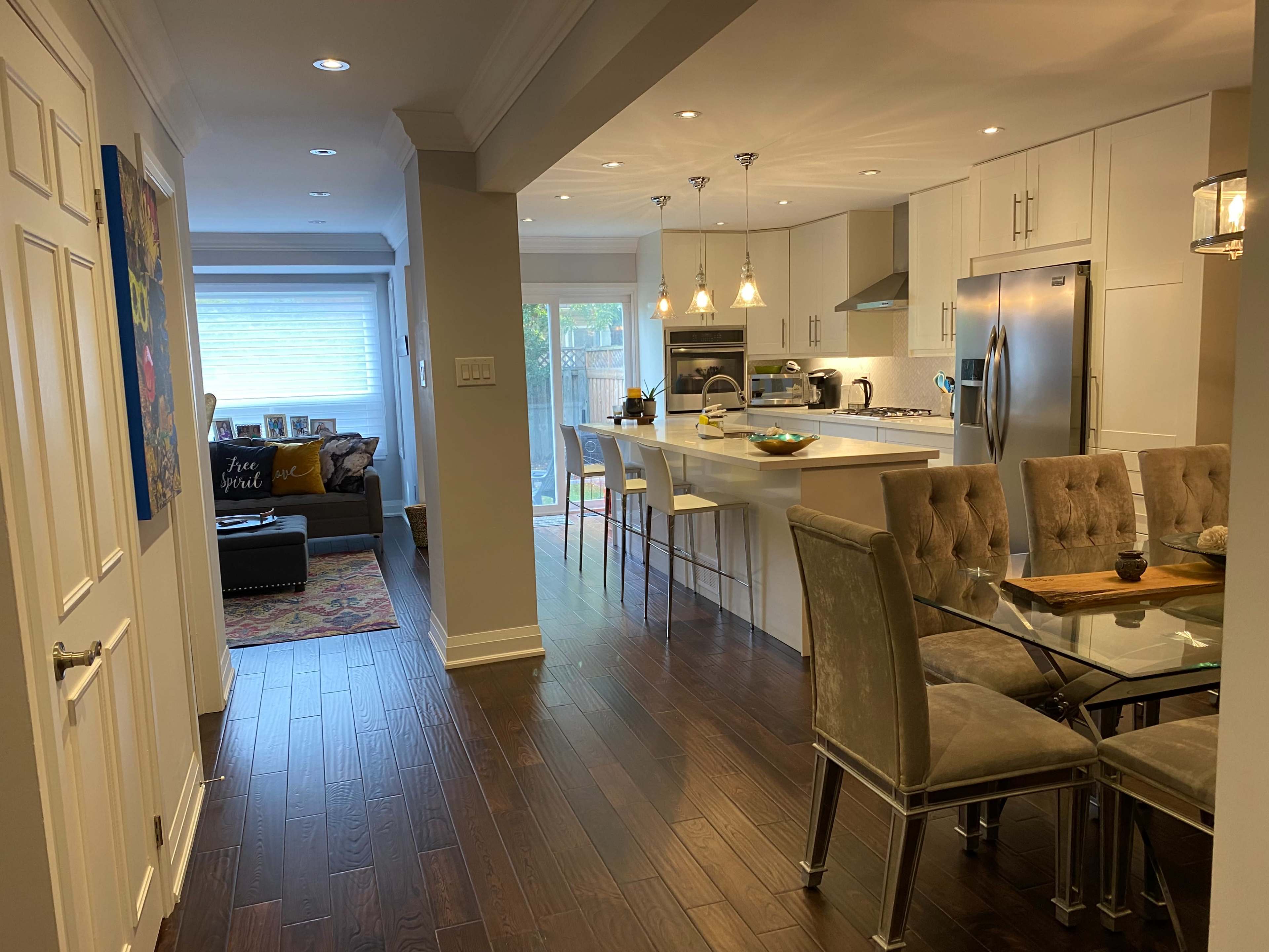 A modern kitchen and dining area with a glass dining table, plush chairs, and a living room visible in the background.