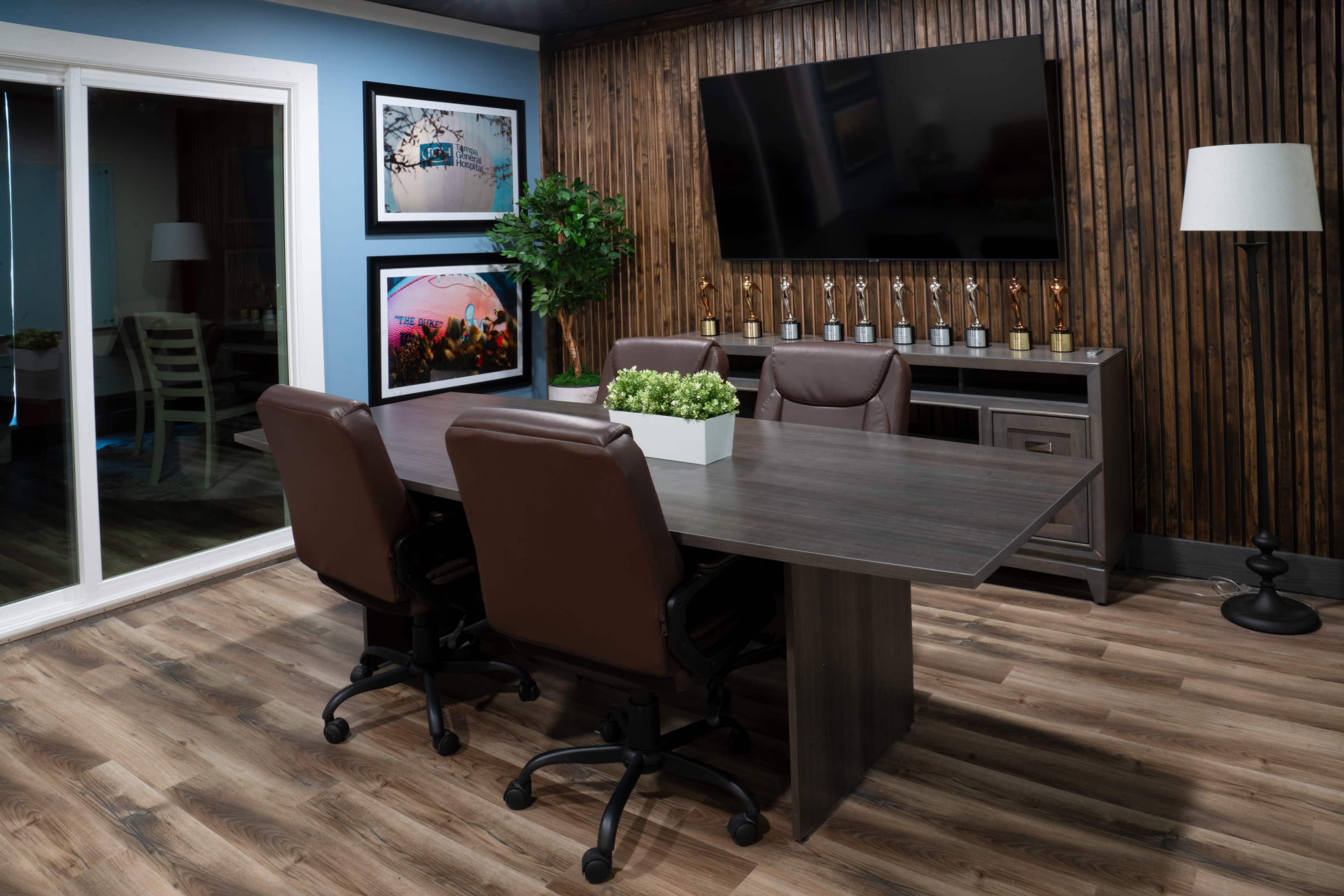 A conference room features a long table with two brown office chairs, a potted plant centerpiece, framed artwork on the walls, and a large wall-mounted TV.