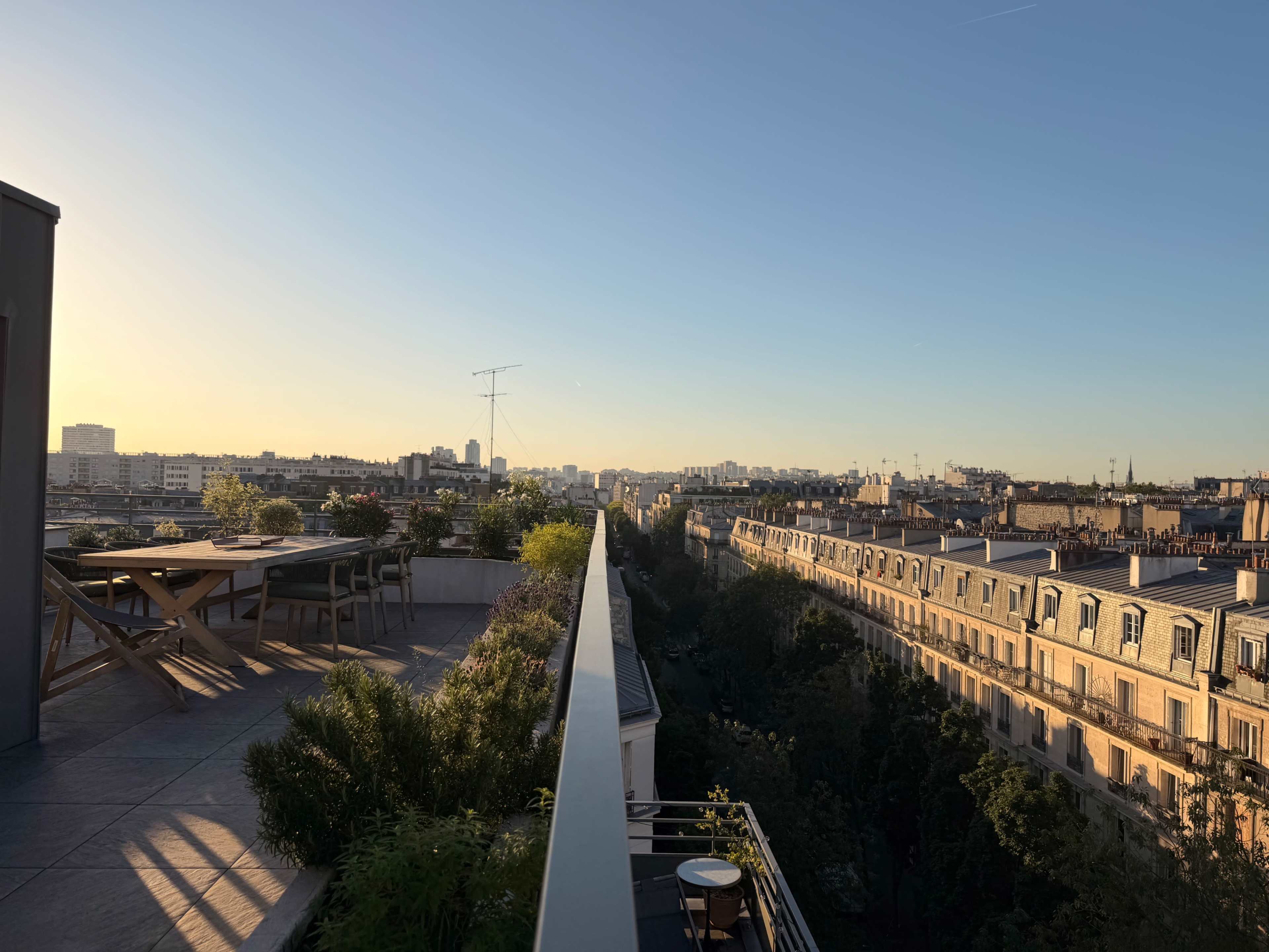 A terrace overlooking a Parisian skyline at sunset, with a long table and chairs on one side and buildings lining the street below.