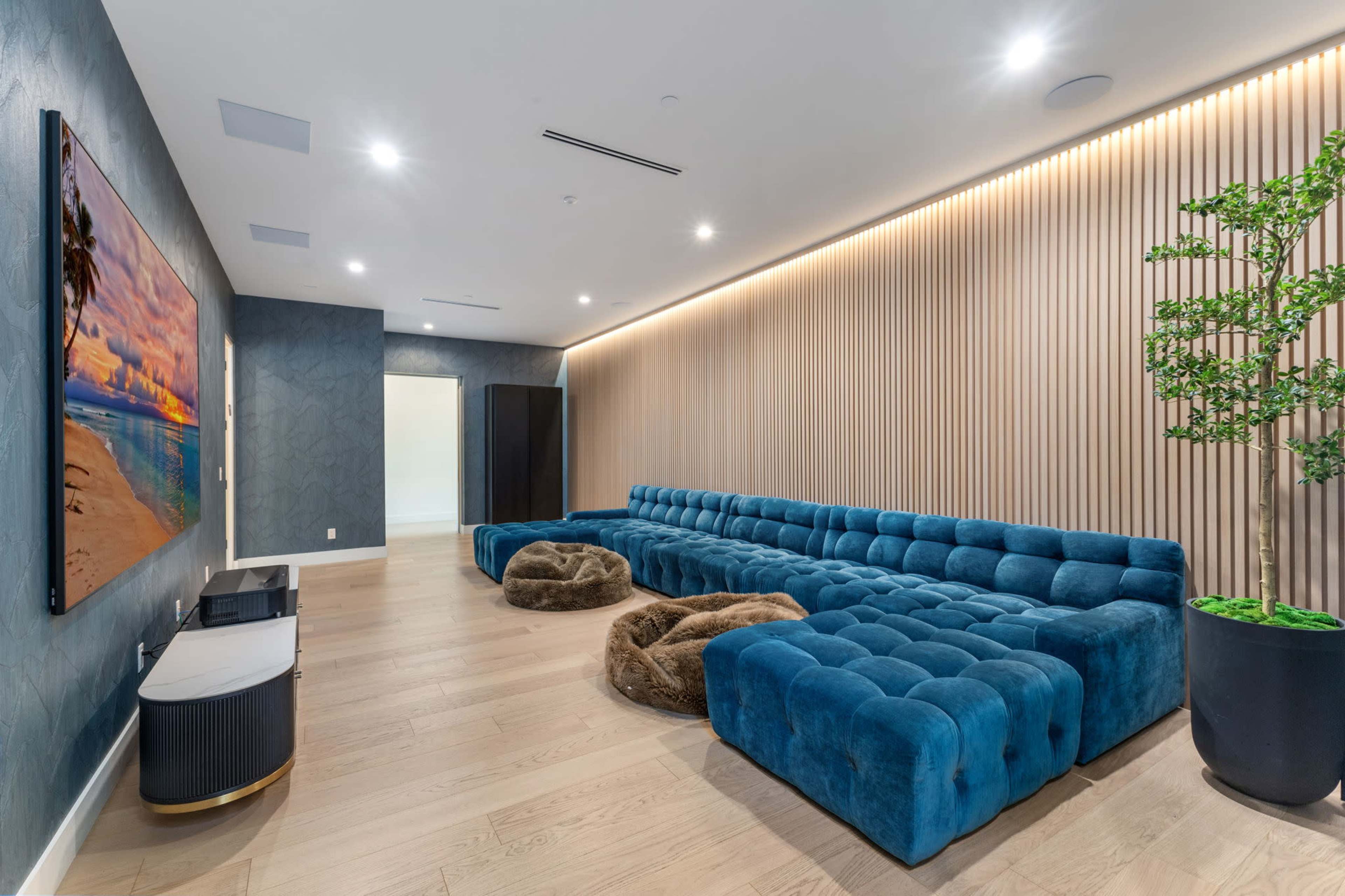 The image shows a modern lounge featuring a large blue tufted sectional sofa, two brown bean bags on the floor, a wall-mounted television, and decorative wall paneling with soft lighting.