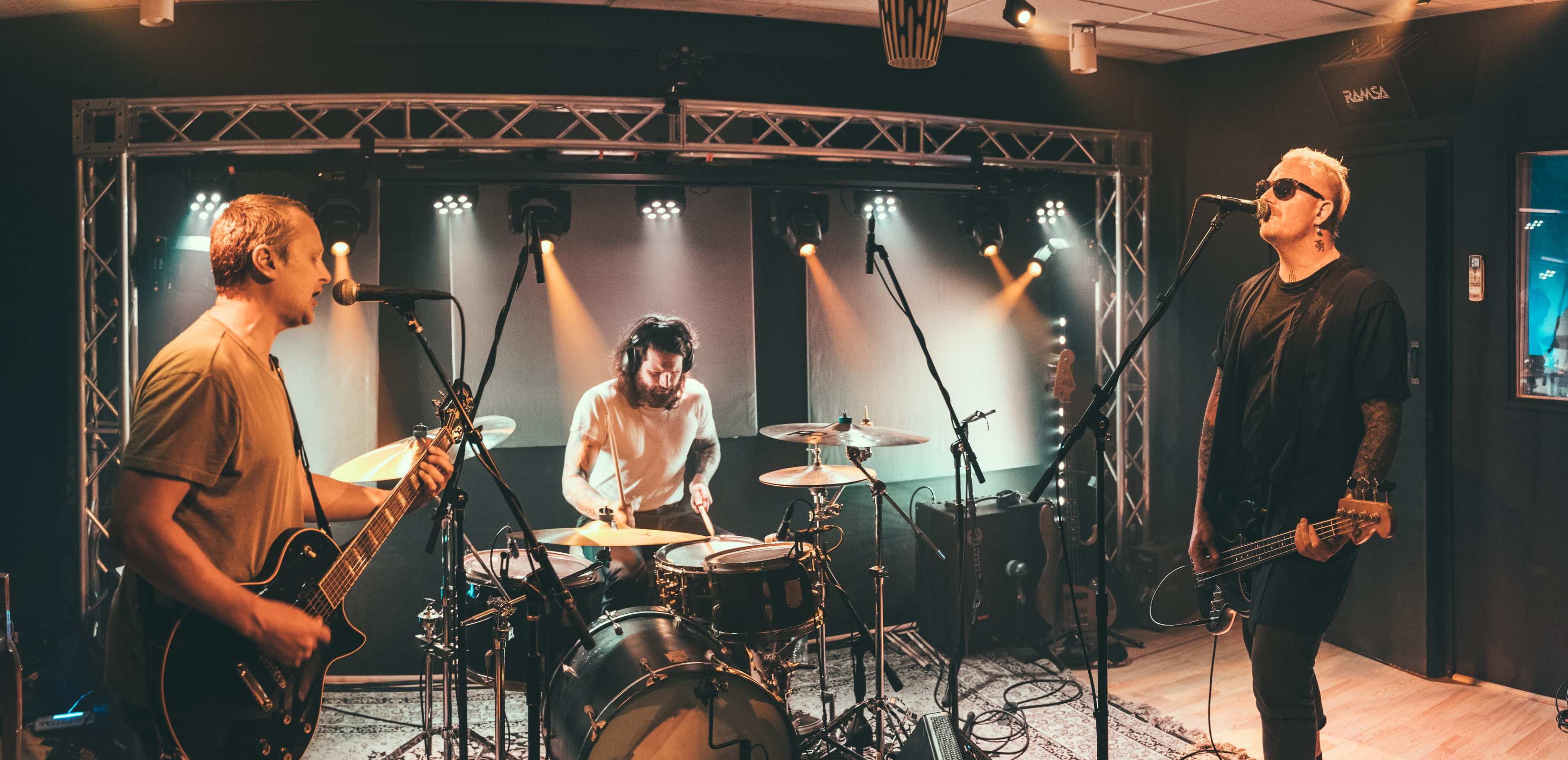 A band performs with a guitar, drums, and vocals in a well-lit studio setting.