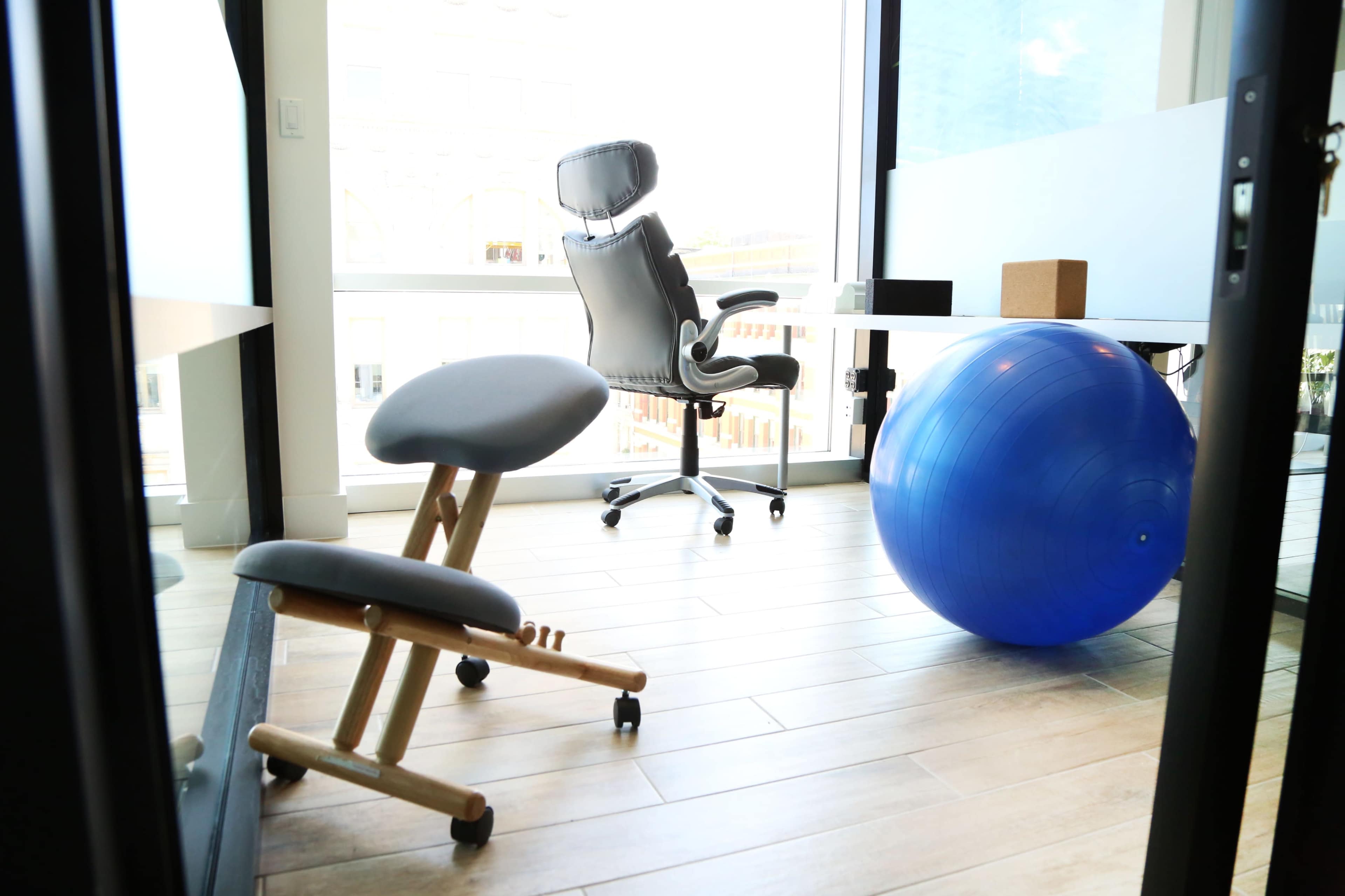 A modern office space featuring a rolling chair, a meditation stool, and a large blue exercise ball near a glass window.