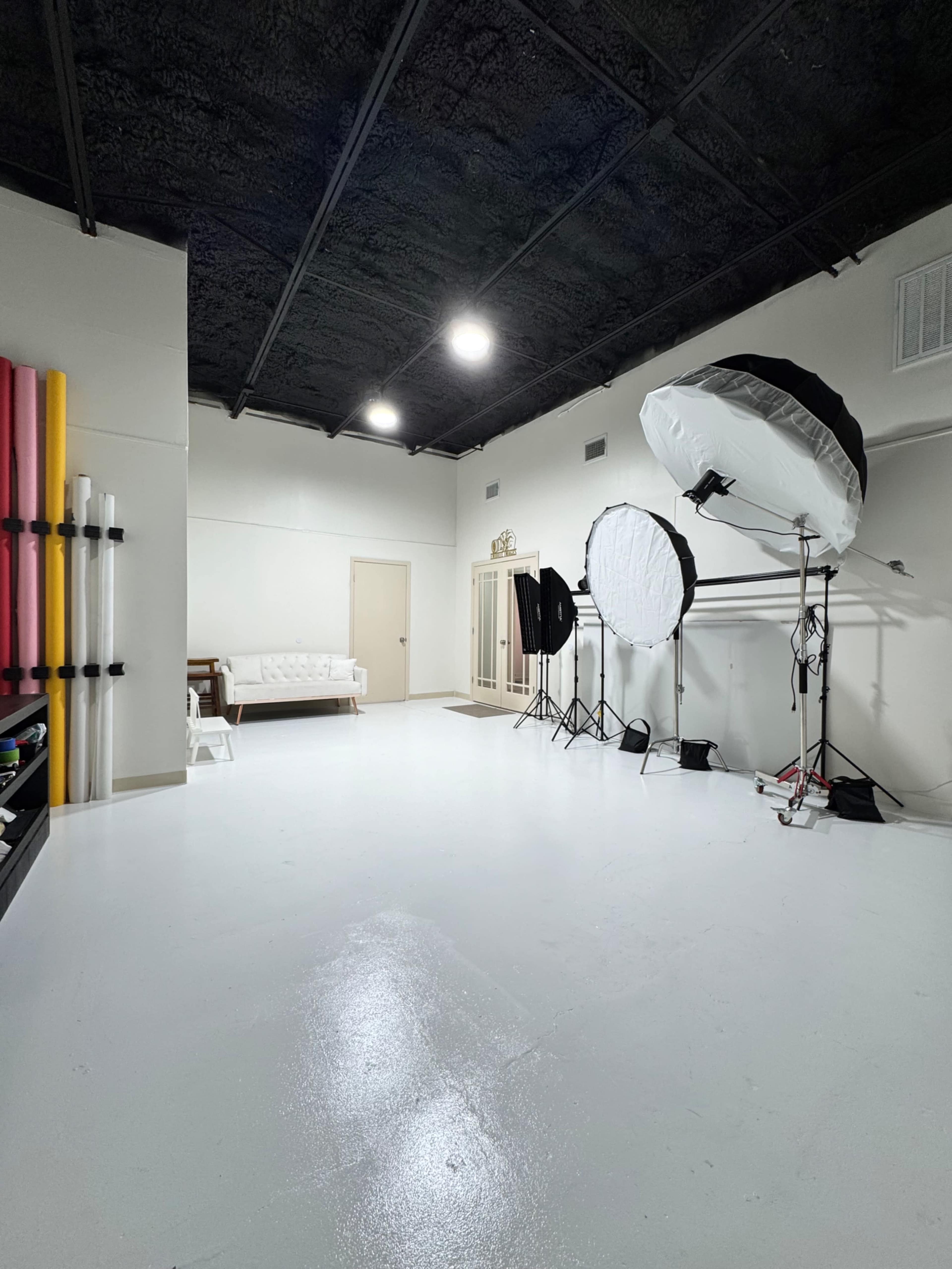 The image shows a photography studio with a white floor, a couch against one wall, and several lighting setups, including large softboxes and backdrop stands.